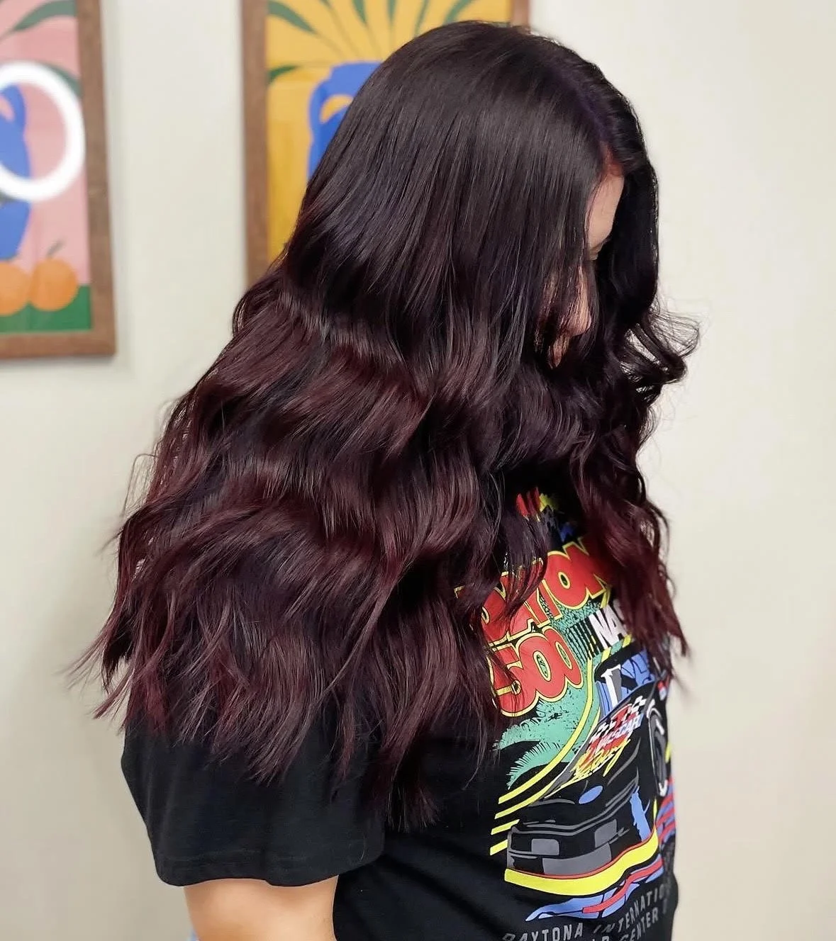 A woman with long, wavy hair dyed in a dark purple shade with a hint of burgundy, wearing a black graphic T-shirt, standing indoors against colorful wall art.