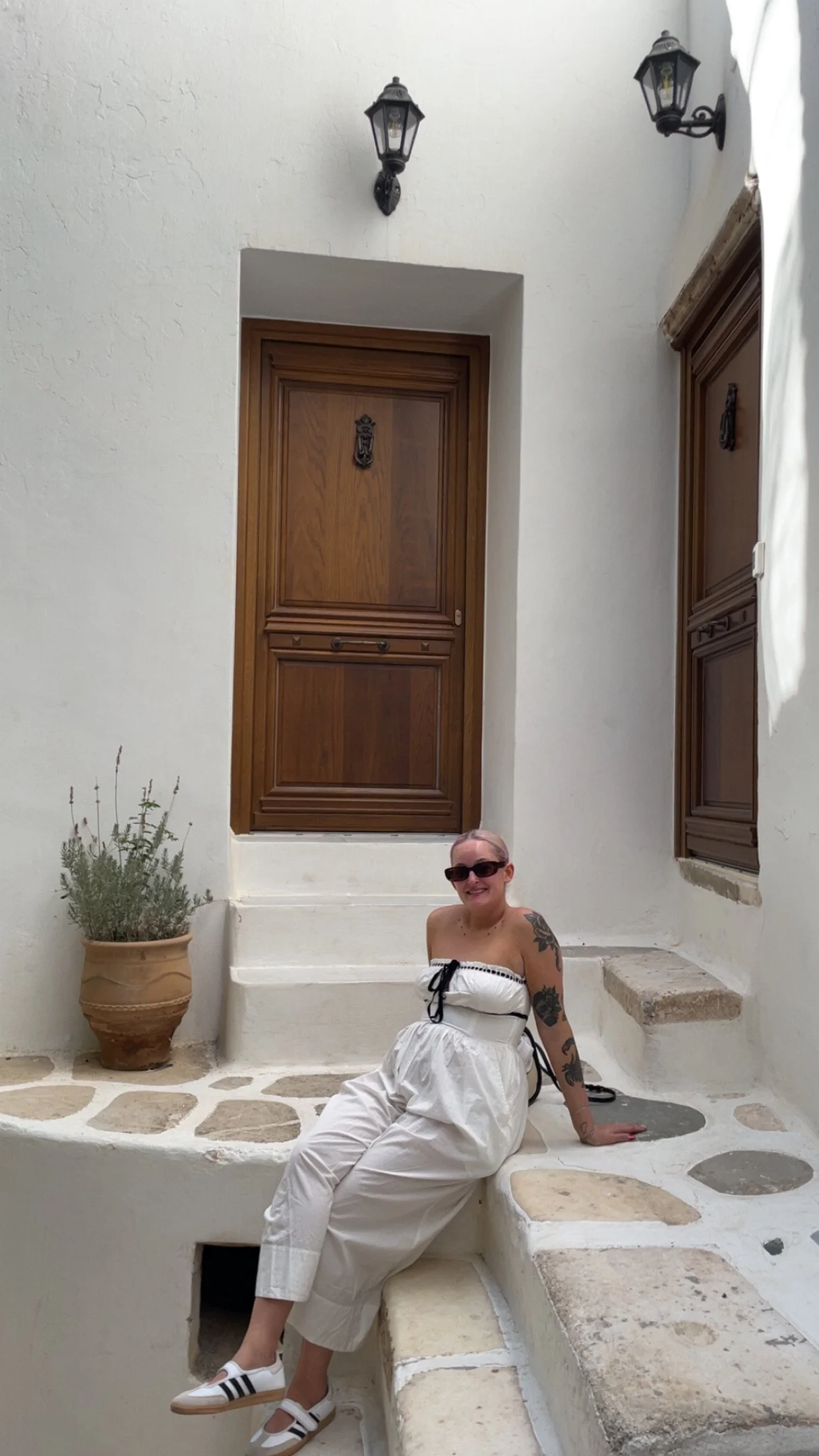 A woman with tattoos, wearing sunglasses and a strapless white dress, sitting on white stone steps outside a building with white walls, wooden doors, a potted plant, and black outdoor wall lanterns.