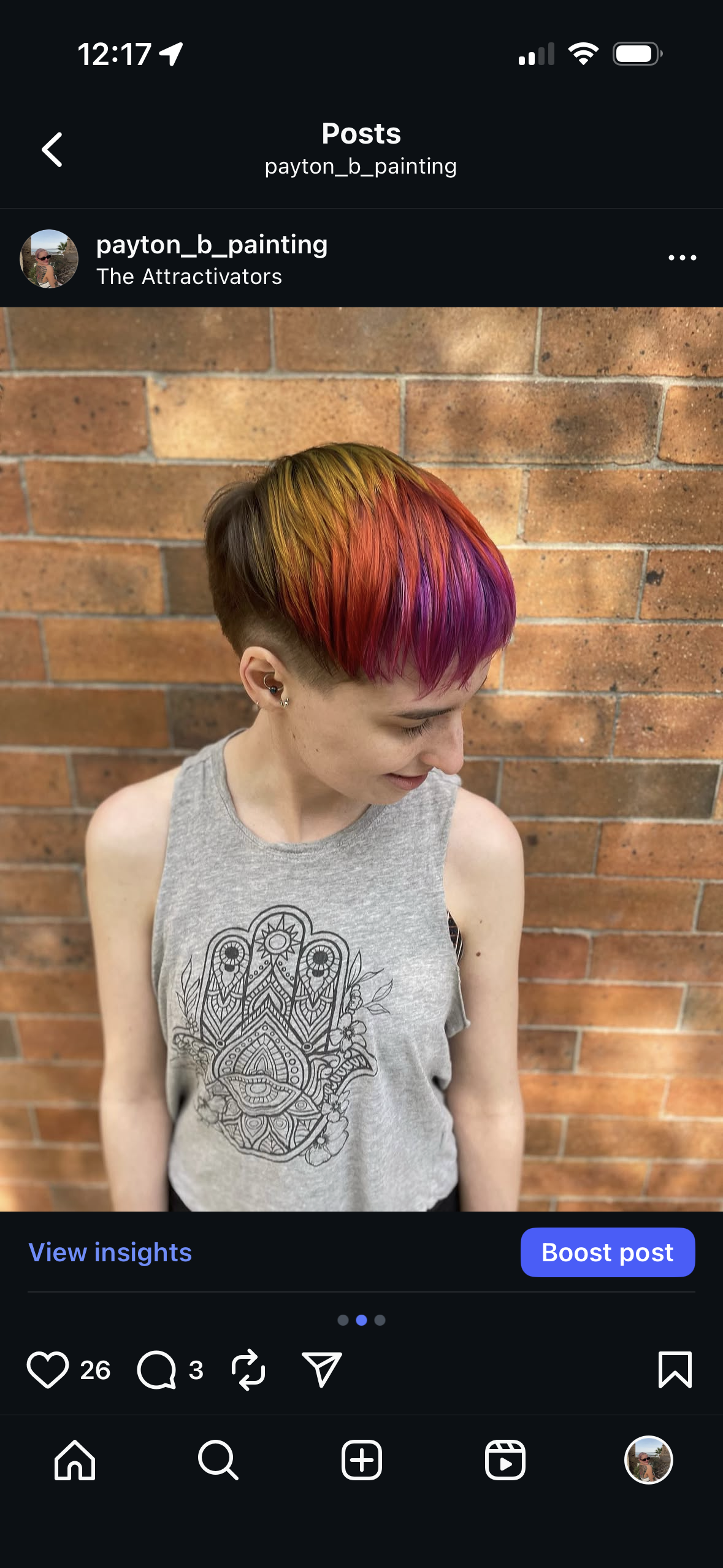A young woman with short hair dyed in rainbow colors, standing against a brick wall, wearing a gray sleeveless shirt with a black intricate design on the front.