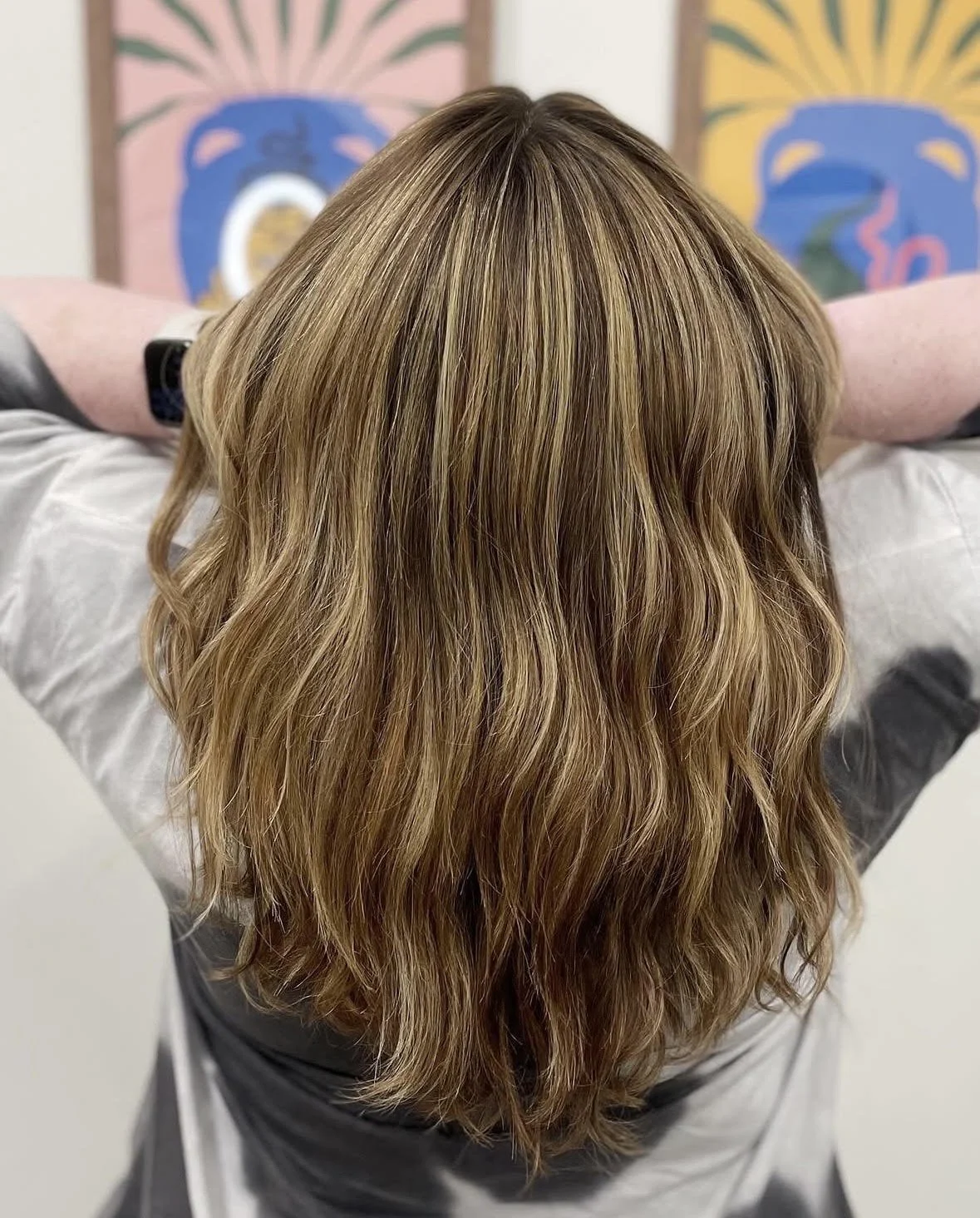 Back view of a person with long, wavy, light brown hair, sitting with hands behind head, with colorful artwork in the background.