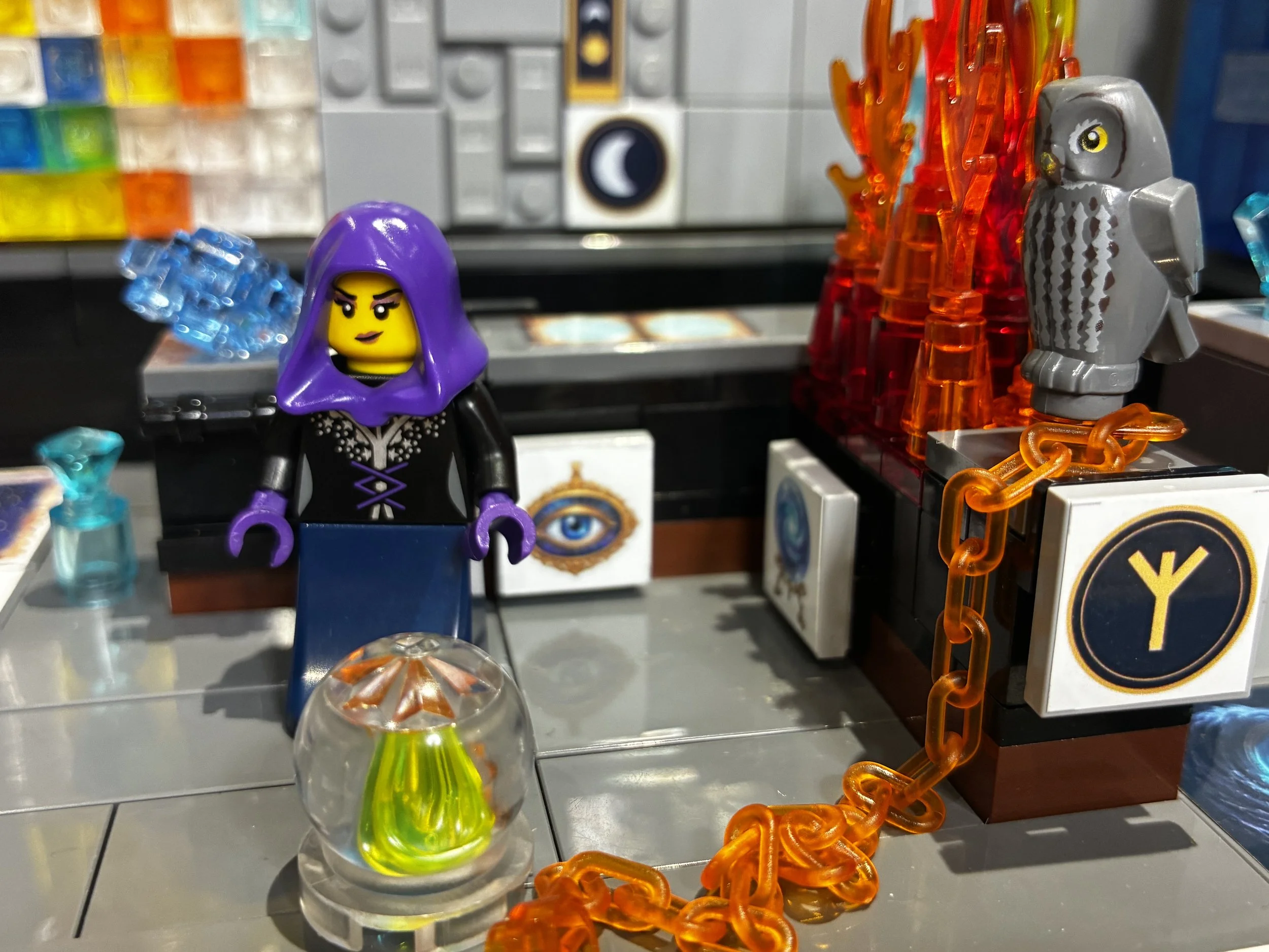 A LEGO wizard tower MOC scene, a hooded seer minifigure and a grey owl both facing a lit crystal ball, dressed with celestial sticker decals.