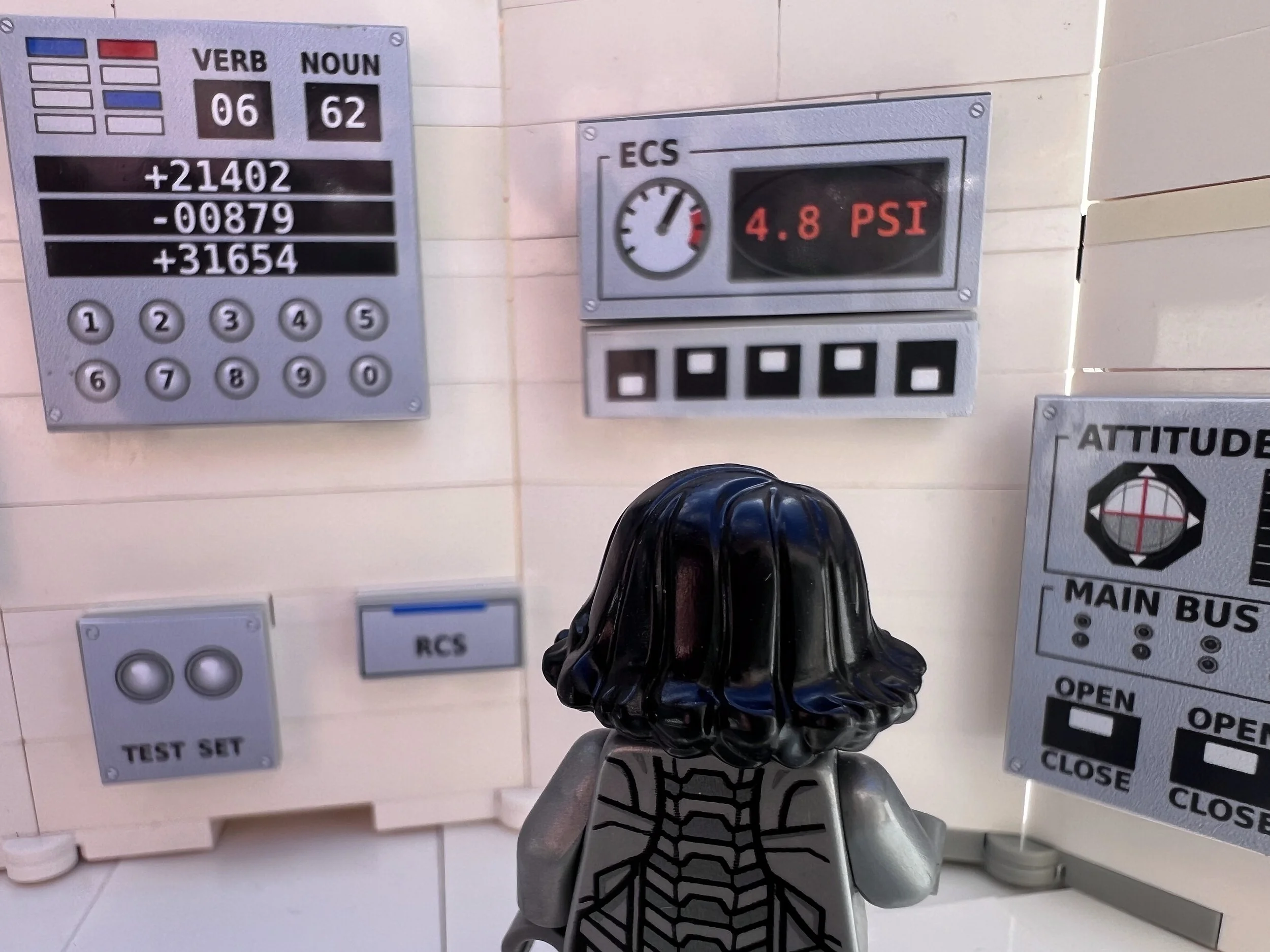 Close view over a minifigure's shoulder of the LEGO guidance computer sticker, VERB 06 and NOUN 62 above three register lines and a number keypad, beside an ECS pressure gauge reading 4.8 PSI.