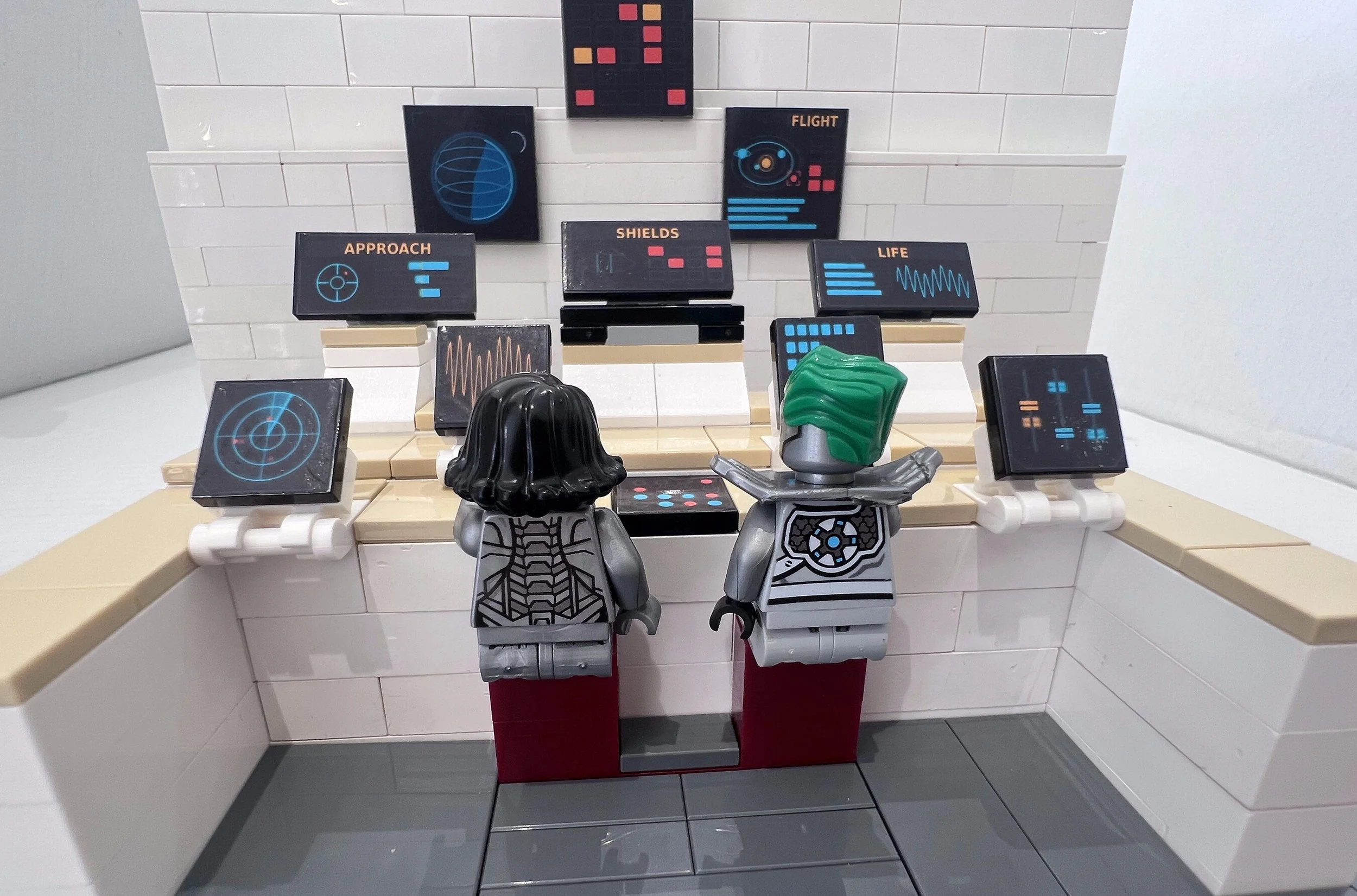 The whole LEGO spaceship bridge MOC, two minifigure crew at their stations facing a wall of screen and readout stickers.