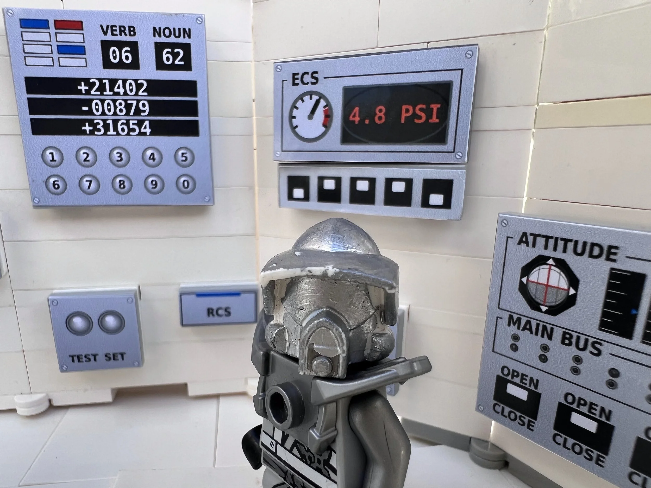 A LEGO minifigure in a silver spacesuit helmet at the retro control console, between the guidance computer, the ECS pressure gauge and an ATTITUDE indicator with MAIN BUS open and close switches.