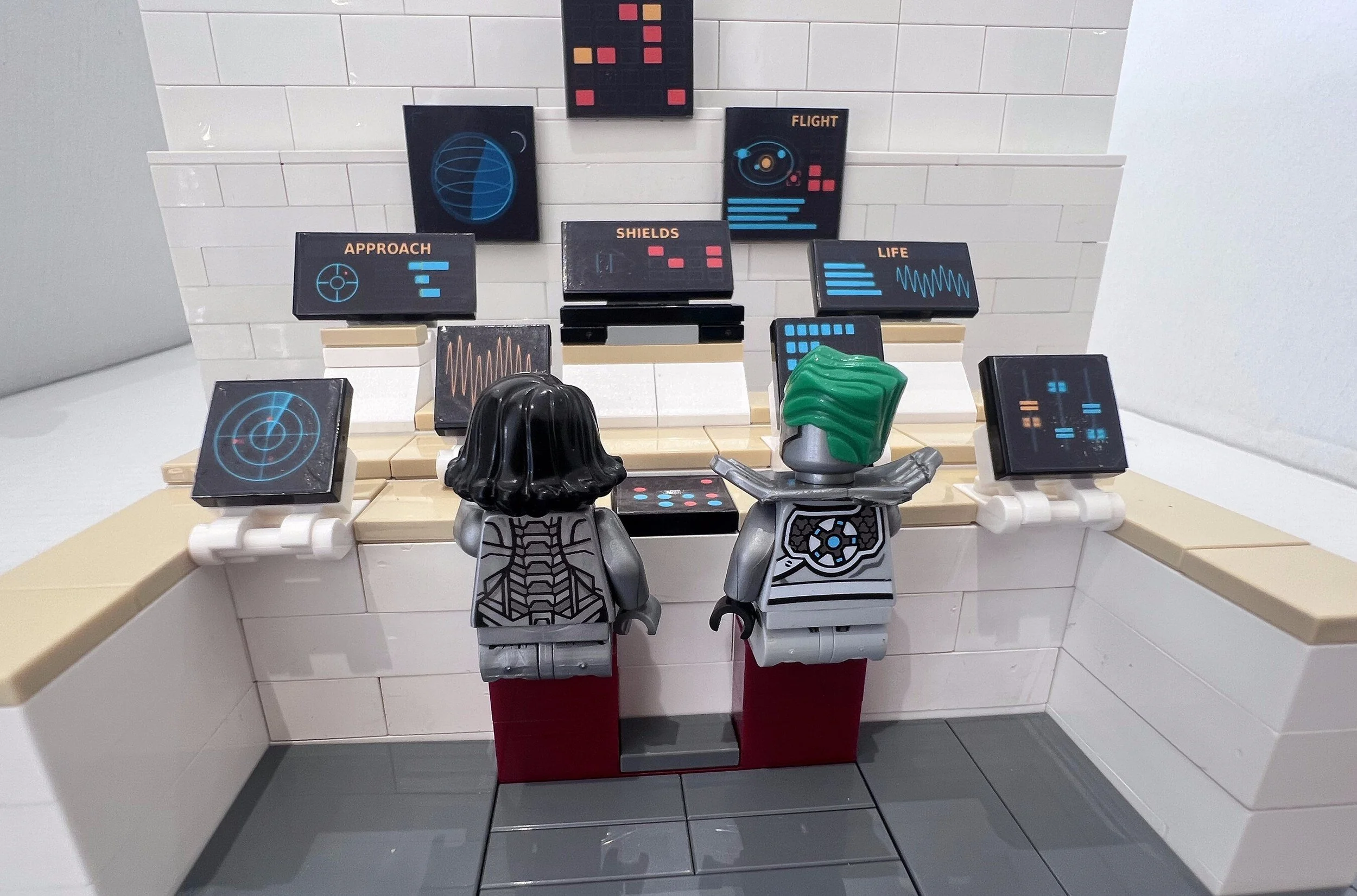 Two minifigure crew at a LEGO spaceship bridge console, with APPROACH, SHIELDS, FLIGHT and LIFE panels on the wall above them