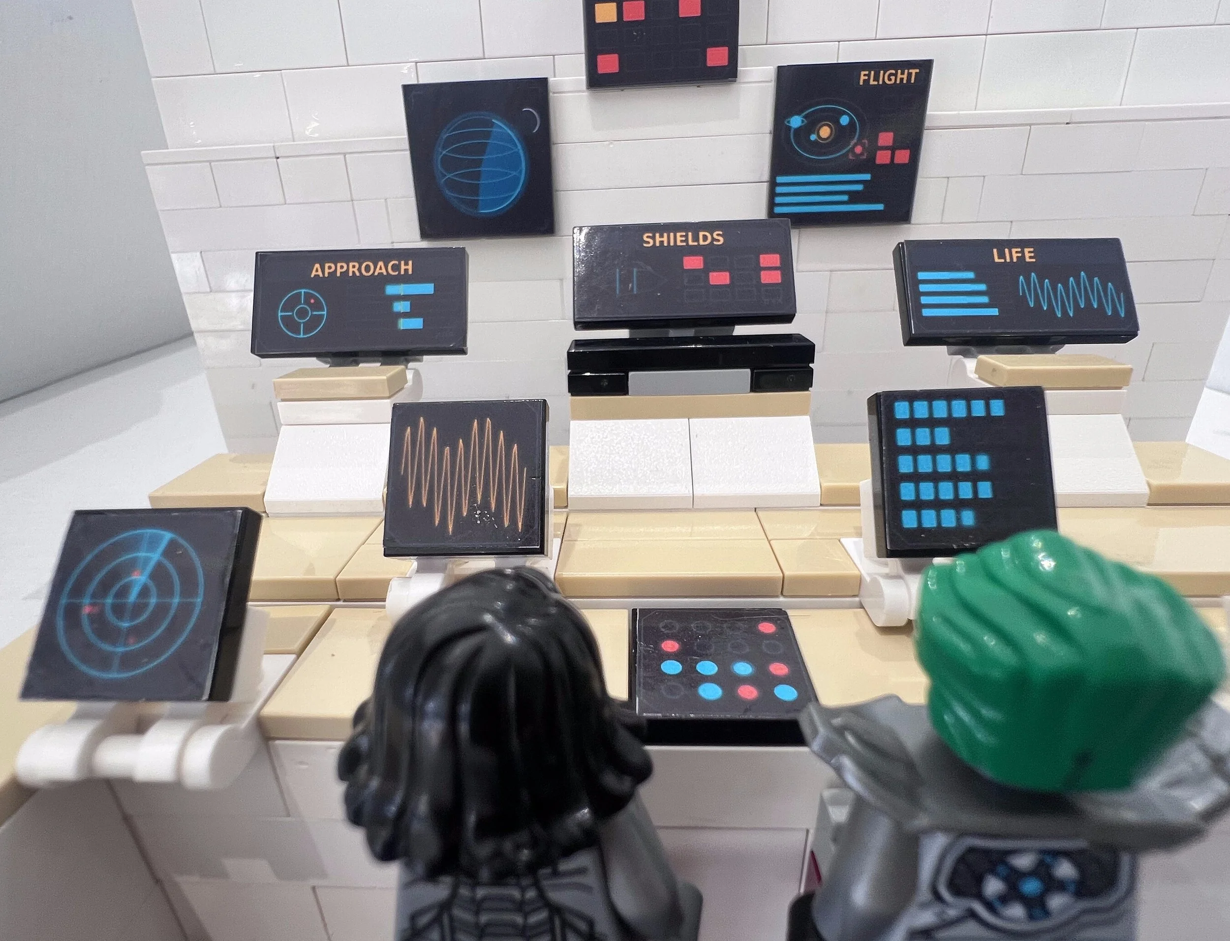 Close view of two LEGO crew stations, each desk tile carrying a different control panel sticker: radar, waveform, keypad and slider readouts.