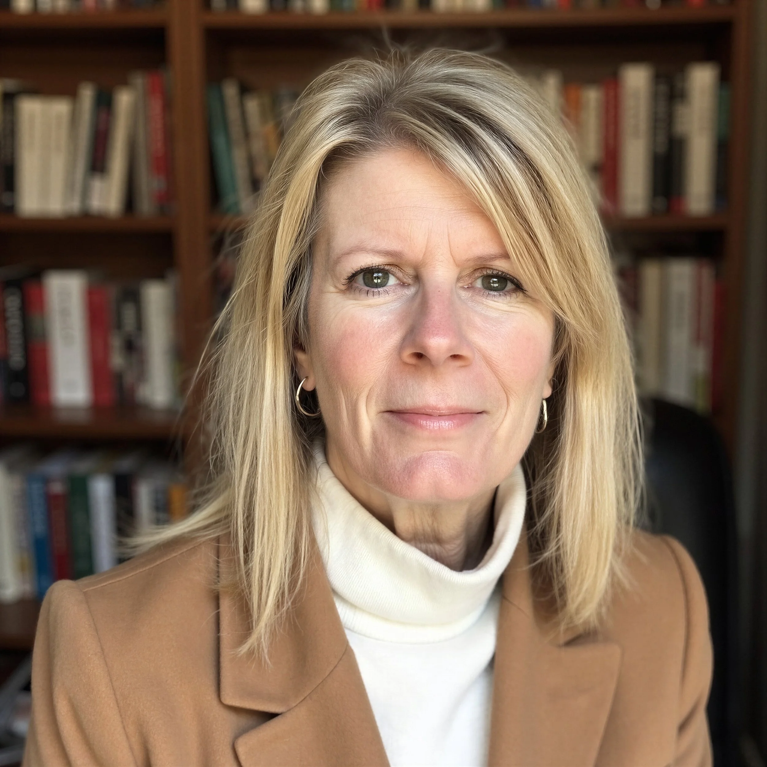 Close-up of a woman with blonde hair, wearing a beige coat and white turtleneck sweater, standing in front of a bookshelf filled with books.
