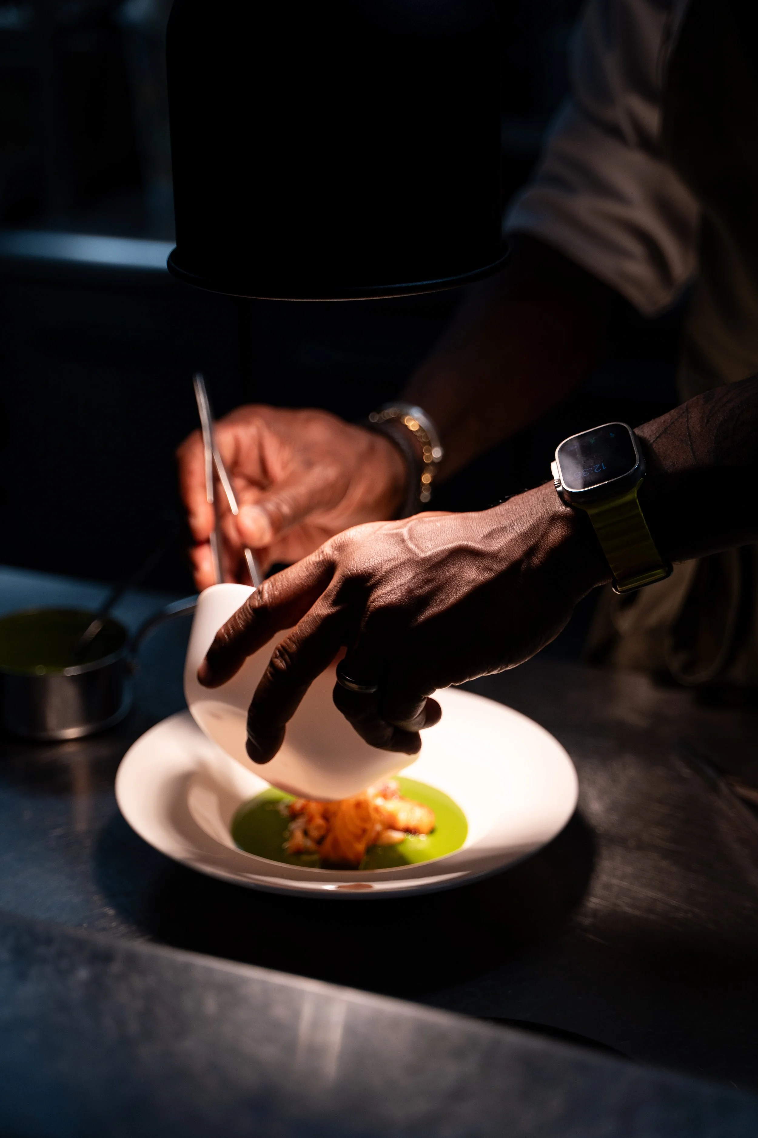 Close-up of a chef plating a dish in a dark kitchen with a black hat in the background, a wristwatch on the chef's left wrist, and a pair of tongs in their right hand, placing food on a white plate with green sauce and vegetables.
