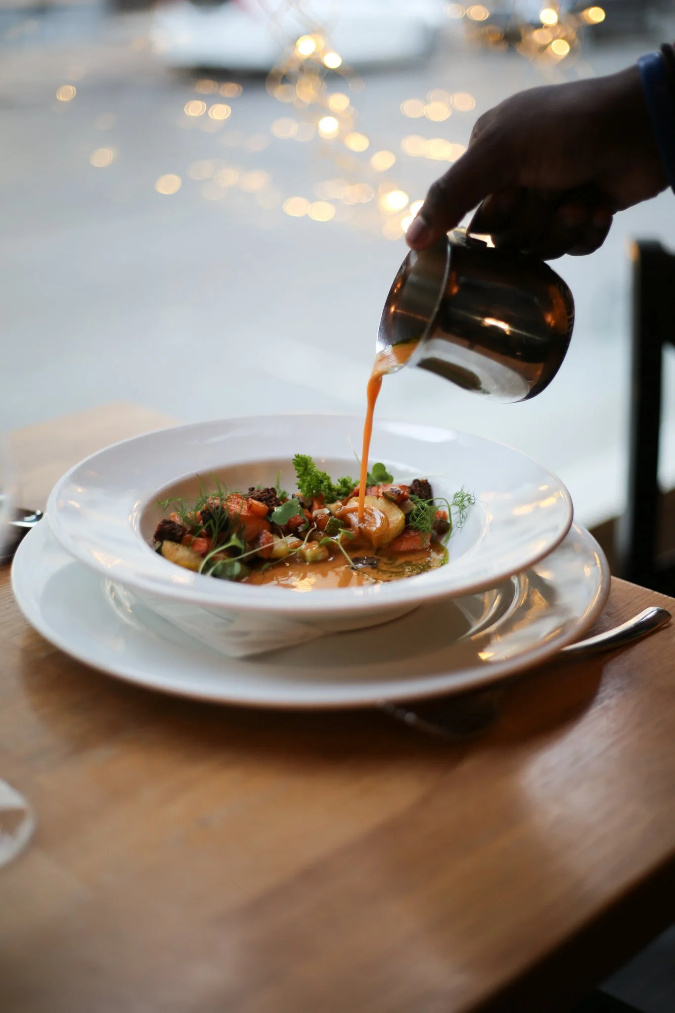 A person pouring sauce over a plated gourmet dish, with a blurred background showing water and lights.