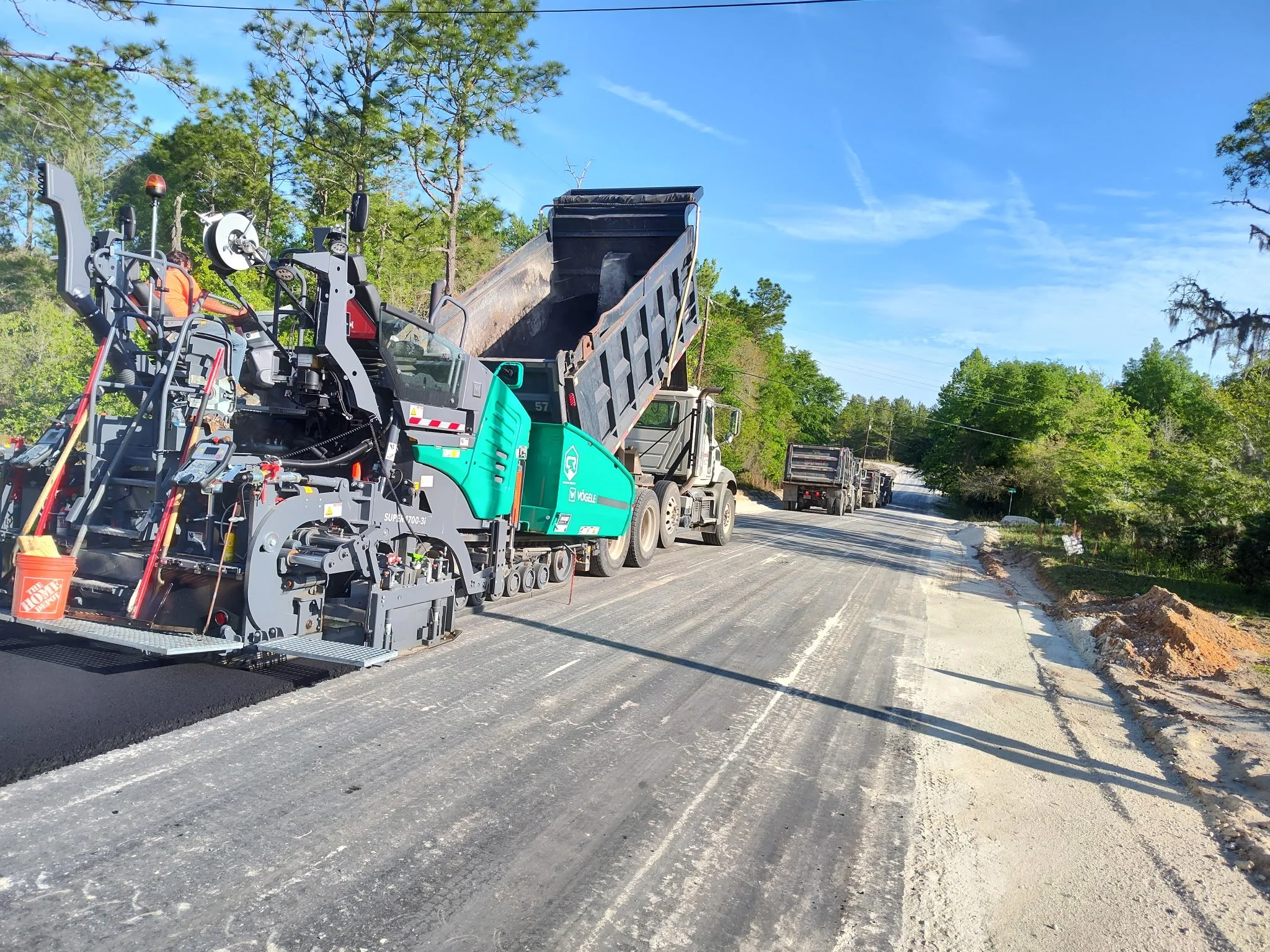 Construction workers operating a large paving machine laying fresh asphalt on a road, with trucks and trees in the background.