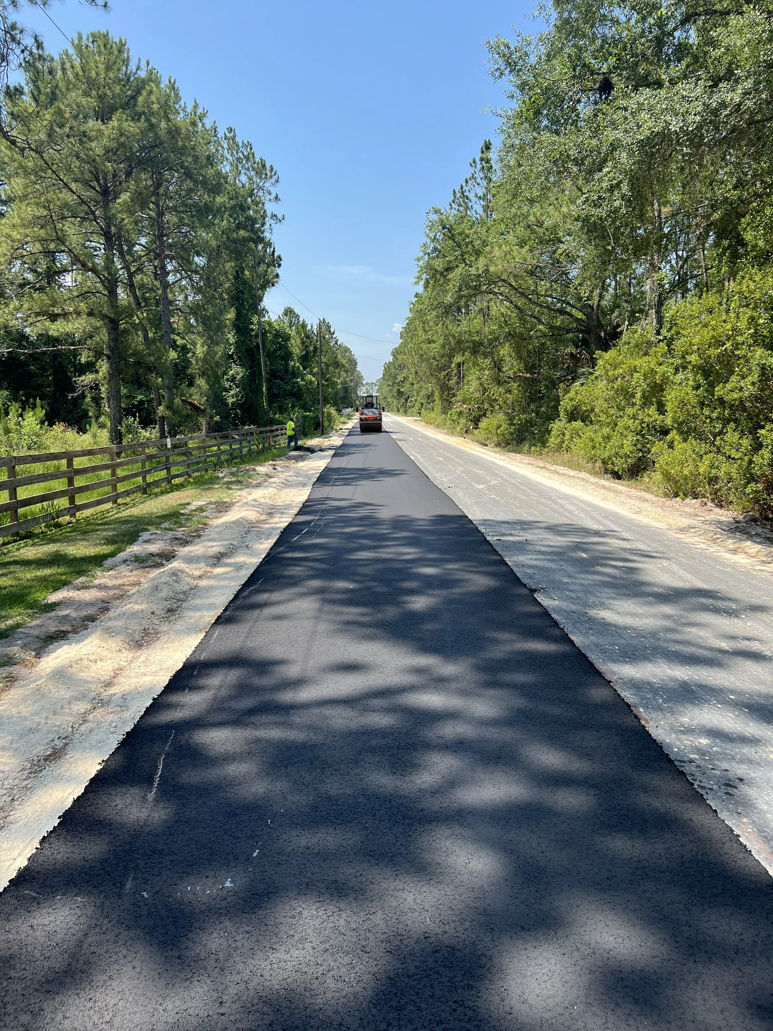 Road construction workers paving a street with asphalt, surrounded by trees and clear blue sky.