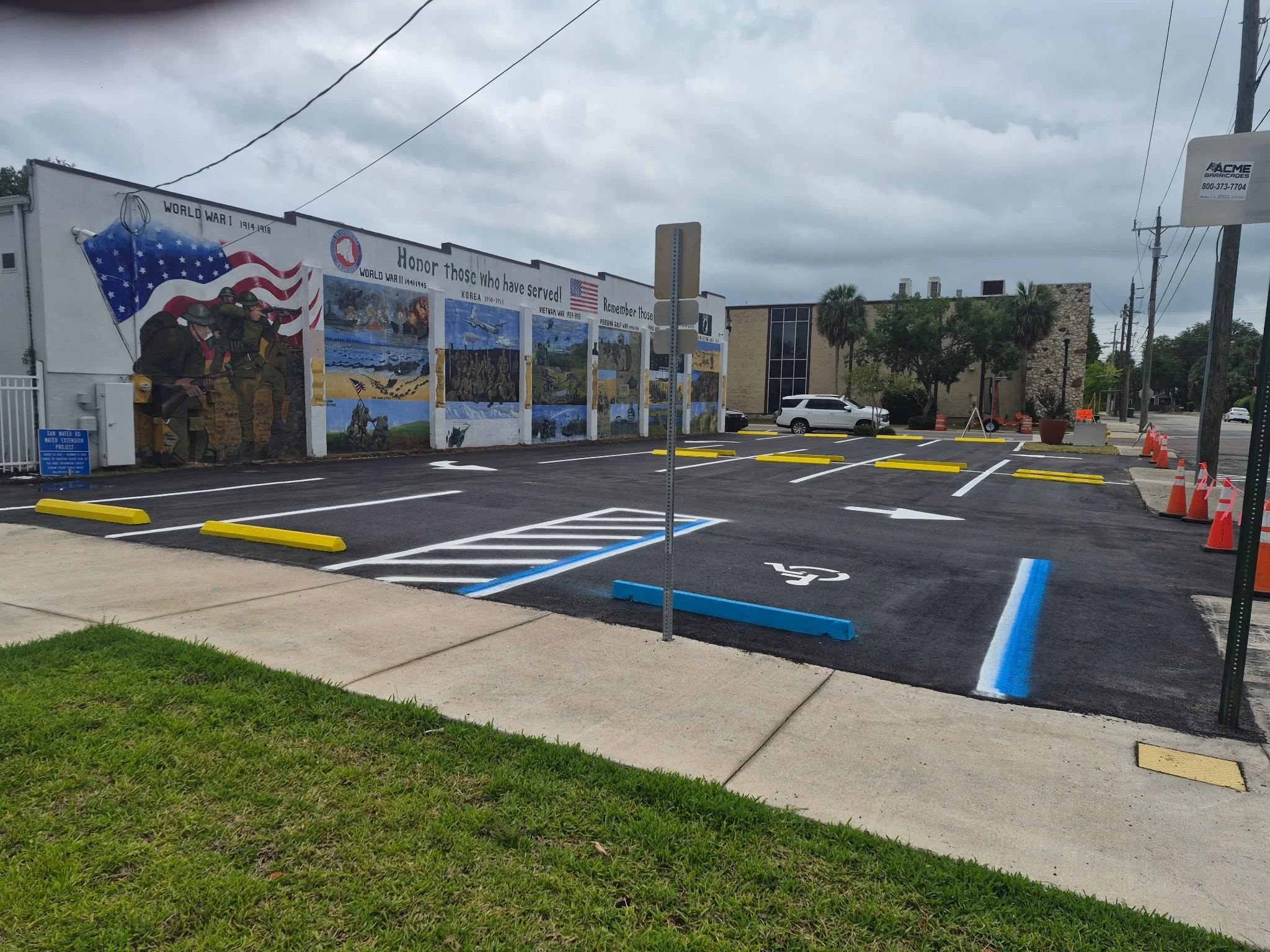 Parking lot under cloudy sky with a mural on the wall honoring military service, featuring soldiers, flags, and scenes from wars; orange traffic cones and a freshly paved and painted parking lot. 