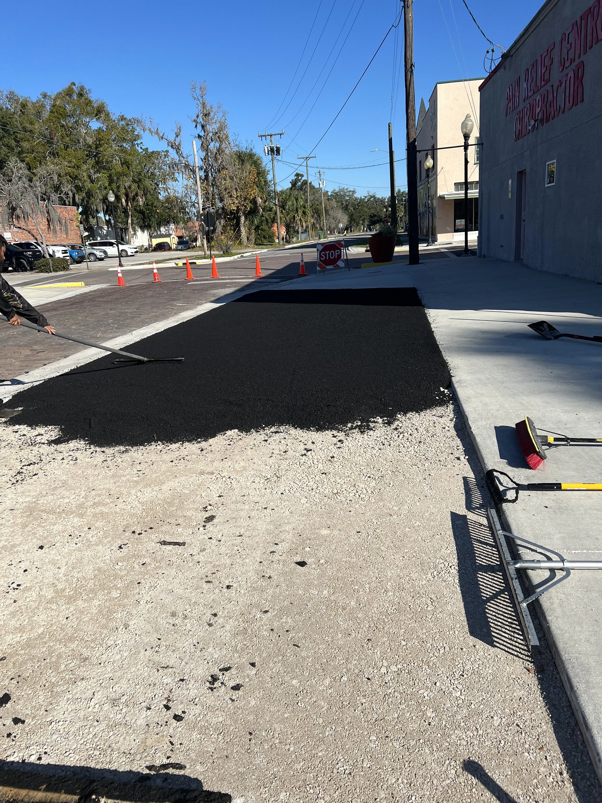 A person is paving a road with black asphalt, and a stop sign and orange cones mark a boundary. The scene takes place in an urban area with parked cars, trees, and buildings in the background.