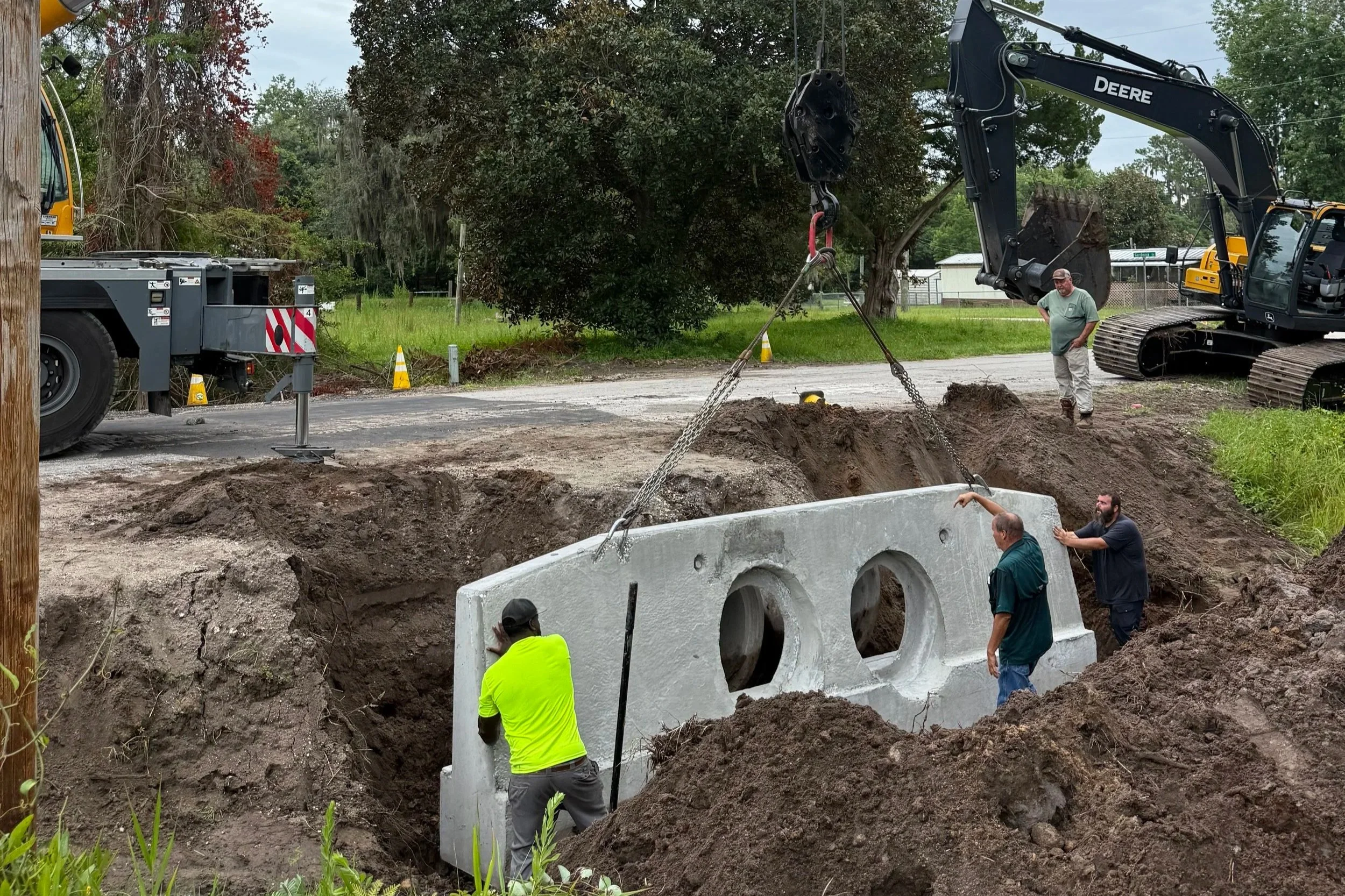 Workers and machinery installing a concrete pipe in a large hole, with trees and residential houses in the background.