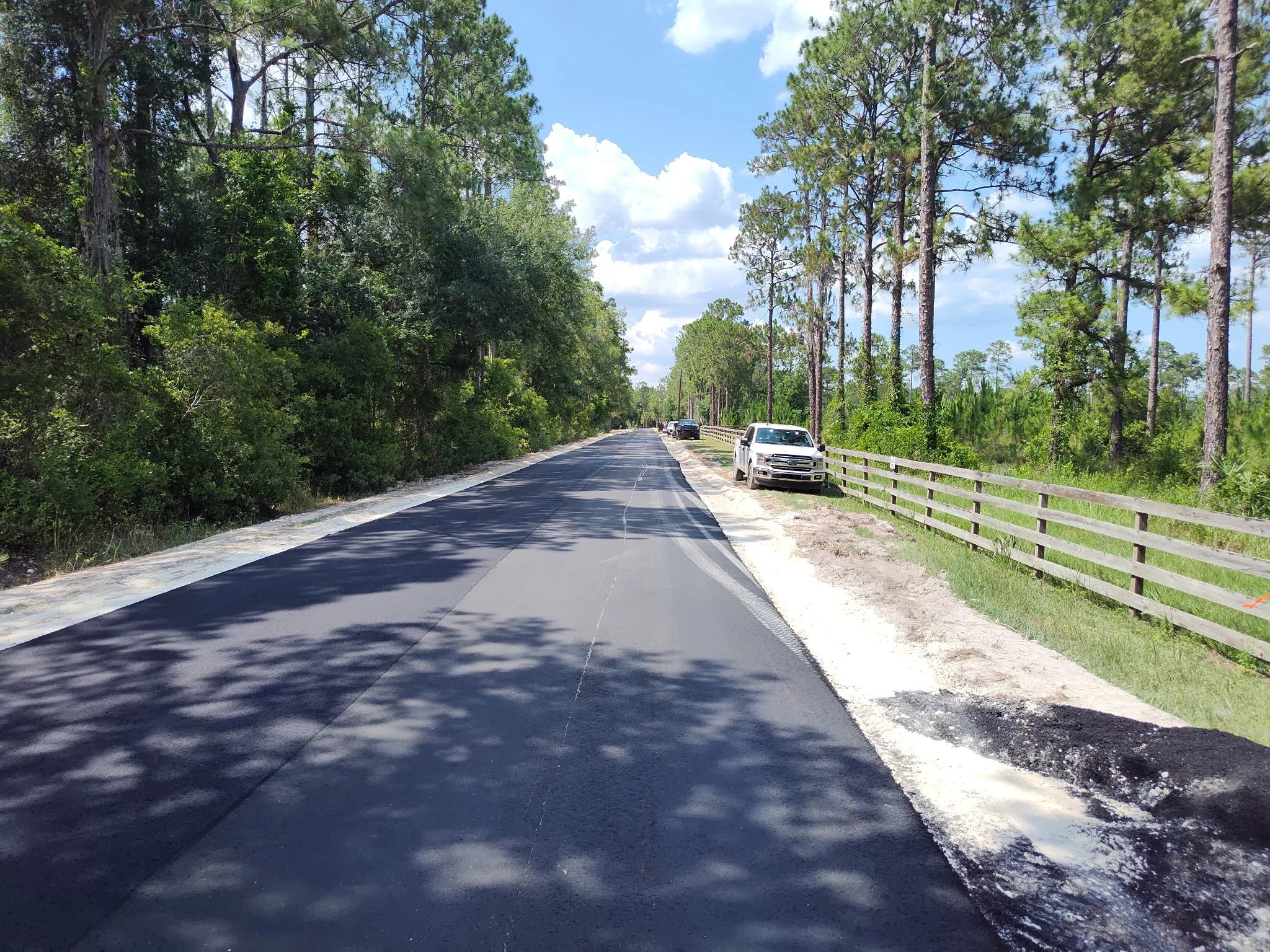 A rural road with freshly paved asphalt, surrounded by tall trees and lush green foliage, with cars parked along the side and a wooden fence on the right.