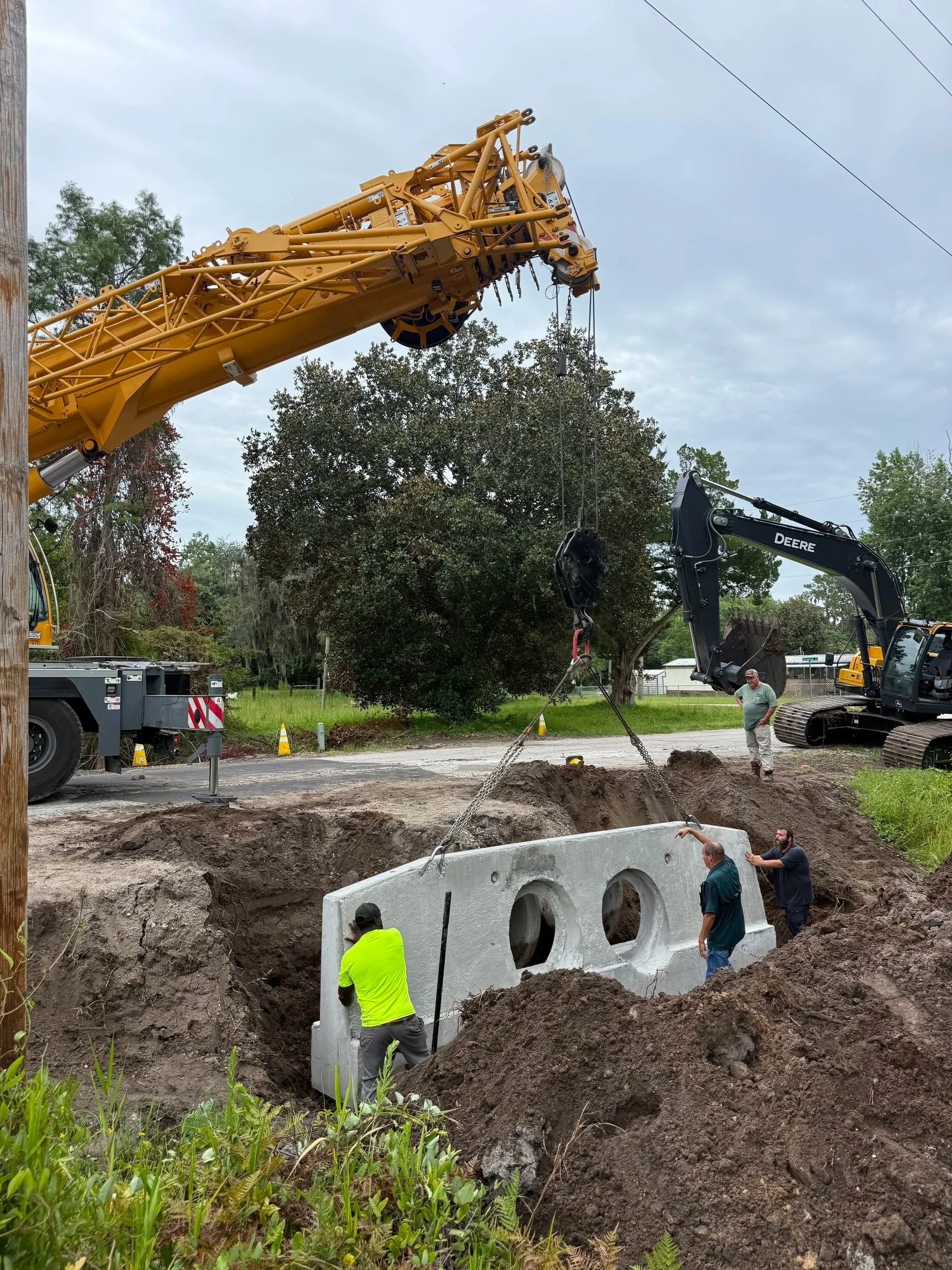 Workers and machinery involved in installing large concrete drainage structure underground during construction project. A crane is placing the concrete piece in the excavated area with several workers guiding and overseeing the process.