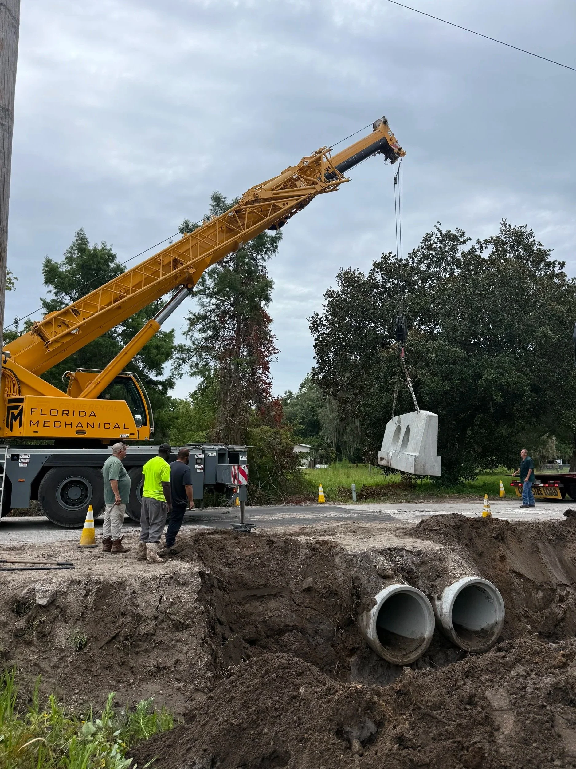 Workers and a crane lifting a concrete block at a construction site with visible underground pipes and exposed soil.