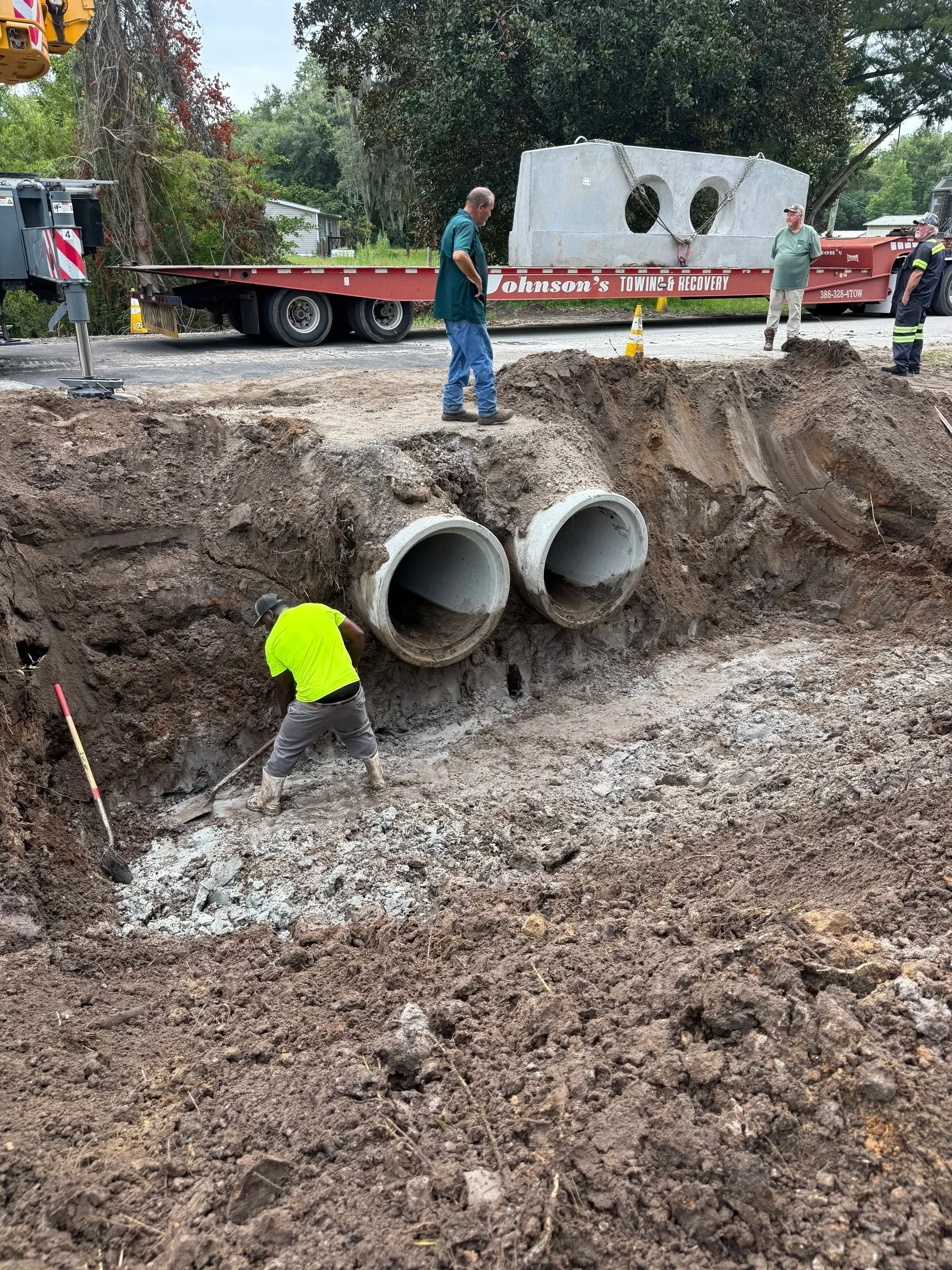 Workers installing underground pipes with large concrete pipe segments and excavation site with dirt and tools, and a flatbed truck in the background.