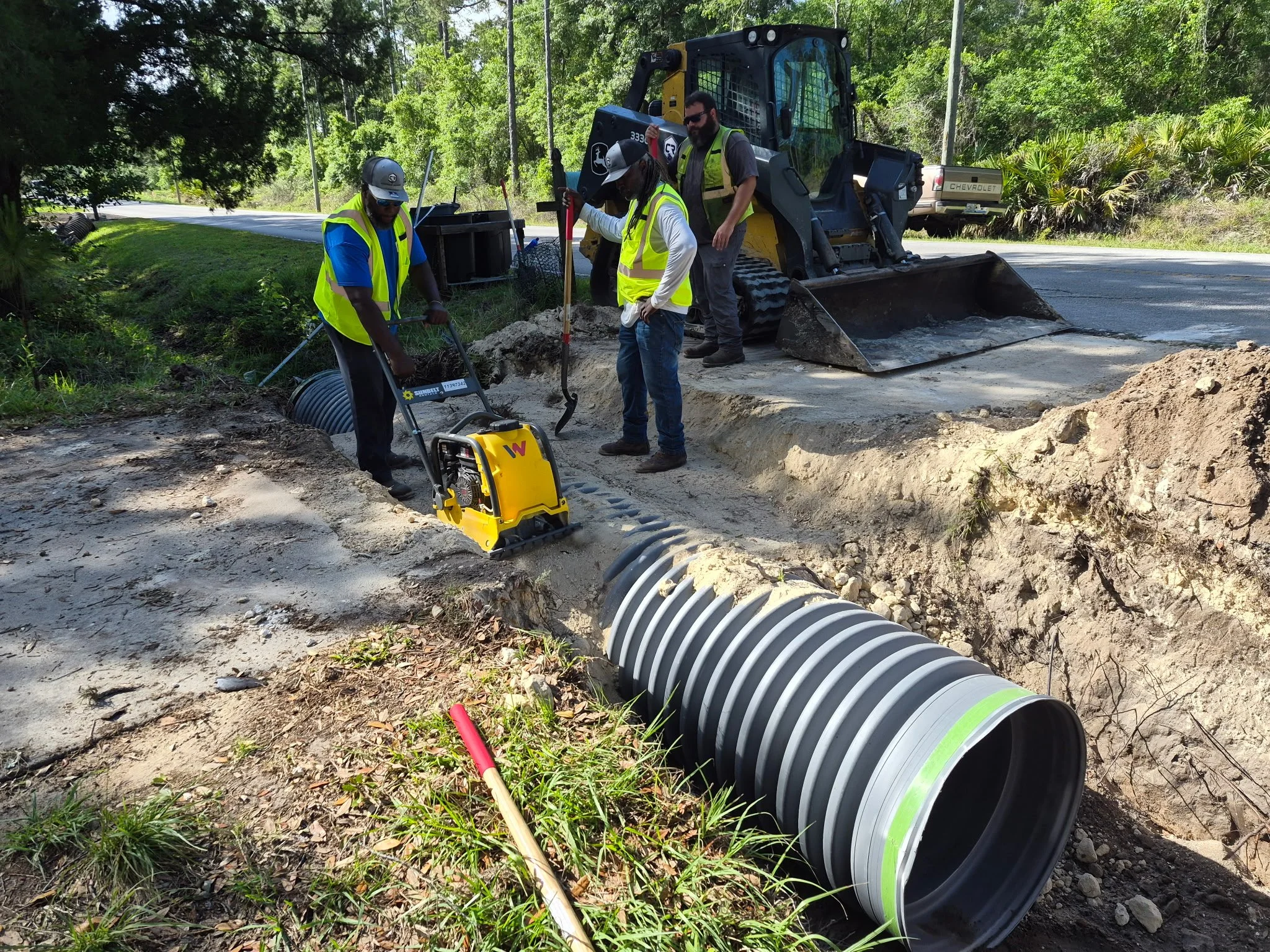 Four workers wearing safety vests and helmets are installing underground drainage pipes along the side of a road. A compact excavator is parked nearby, and the workers are handling a large black corrugated pipe.