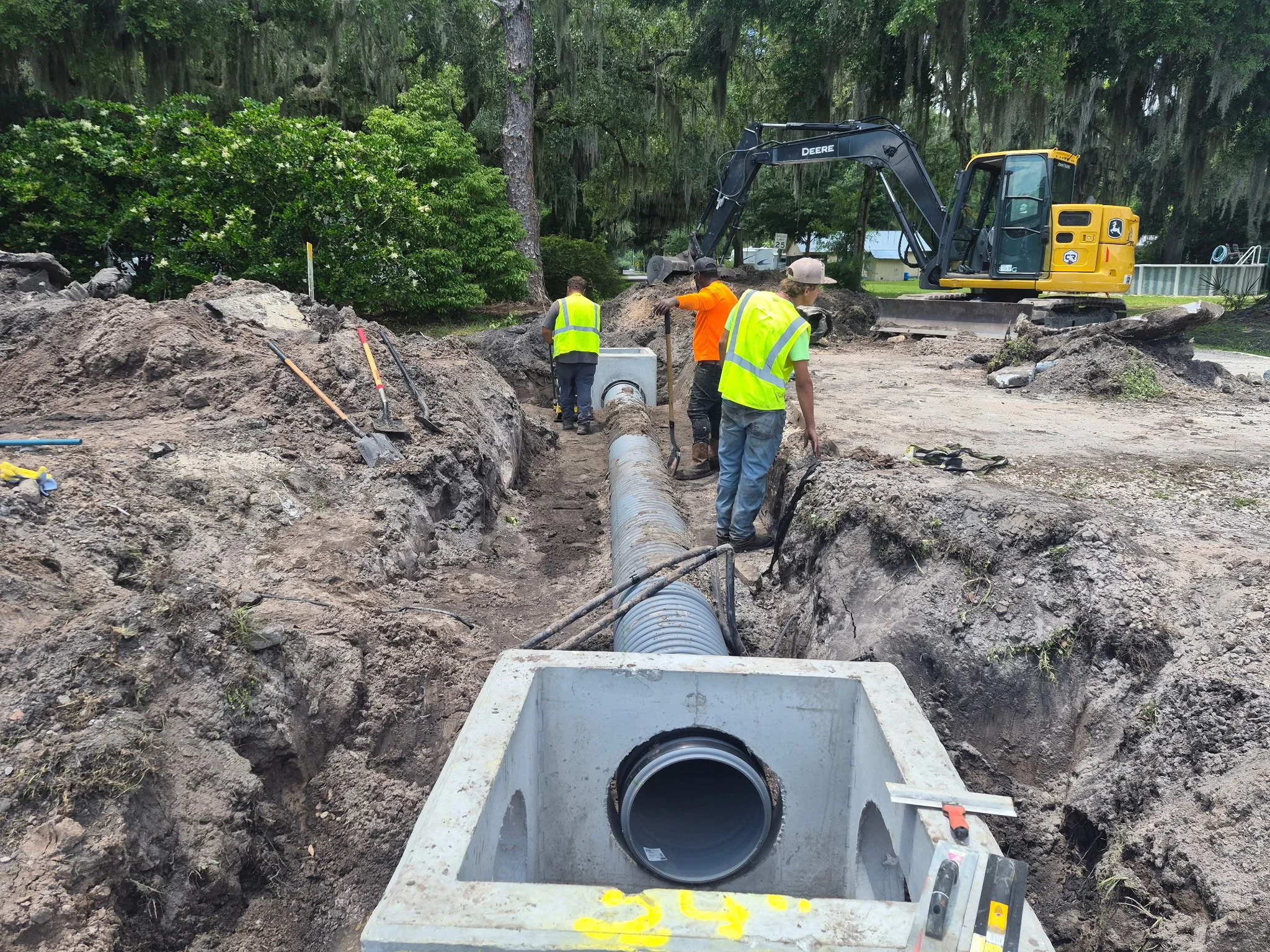 Construction workers operating machinery and installing underground pipes in a residential outdoor area surrounded by trees and greenery.