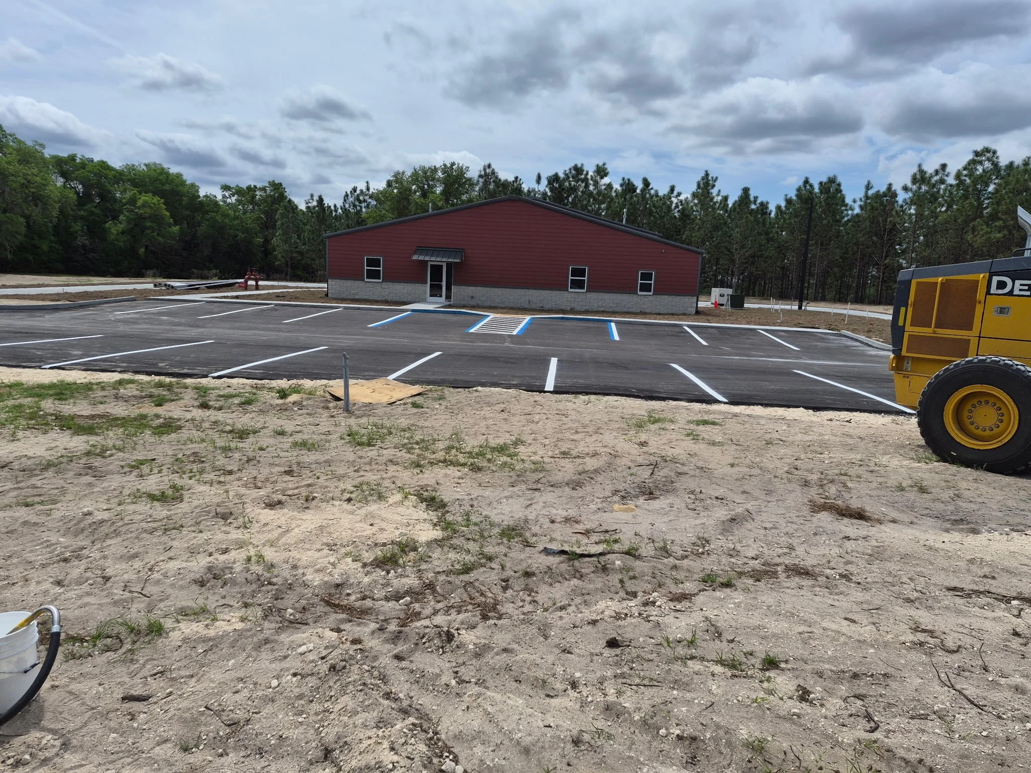 A newly constructed building with red siding and a gray stone foundation, surrounded by freshly paved and painted parking spaces on a parking lot, with construction equipment visible in the foreground, and a background of trees and cloudy sky.