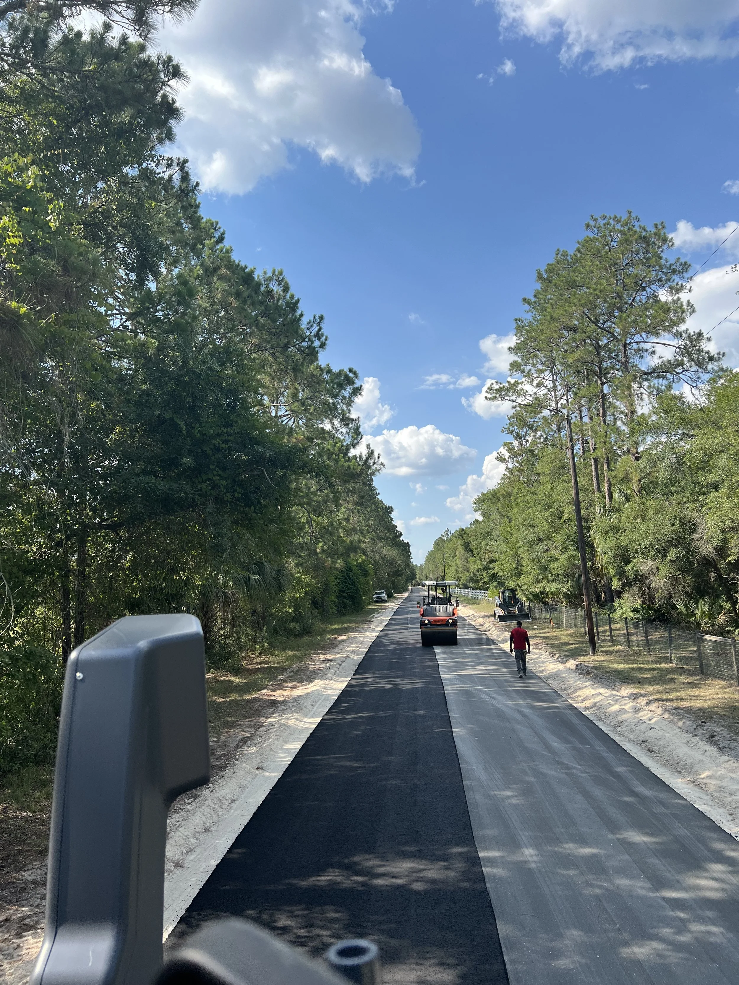 Road construction scene with a partially paved road, surrounded by trees on both sides and a bright blue sky with some clouds. Construction vehicles and workers are present on the road.