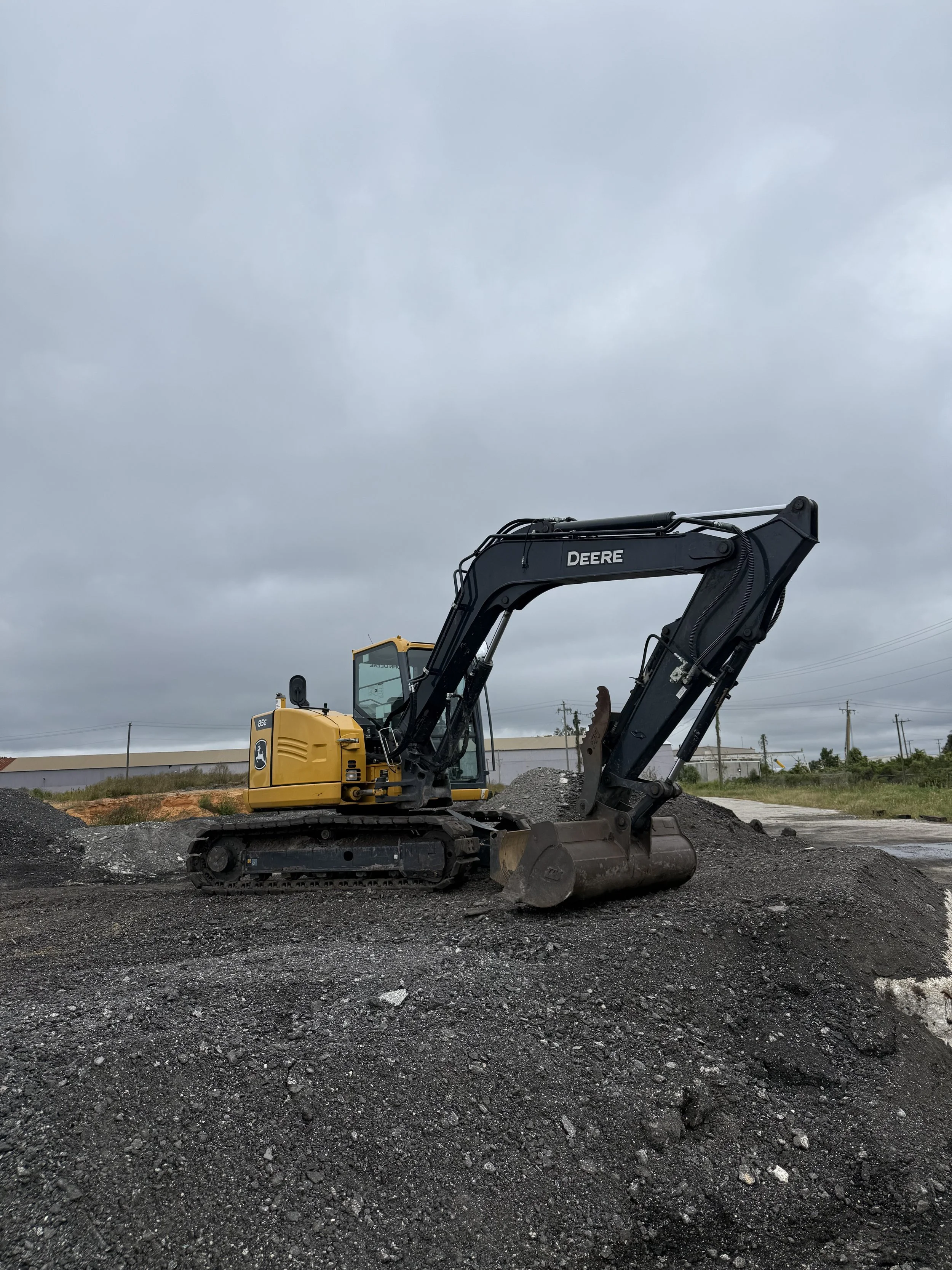 A yellow and black John Deere excavator working on a construction site with dark asphalt millings and an overcast sky.