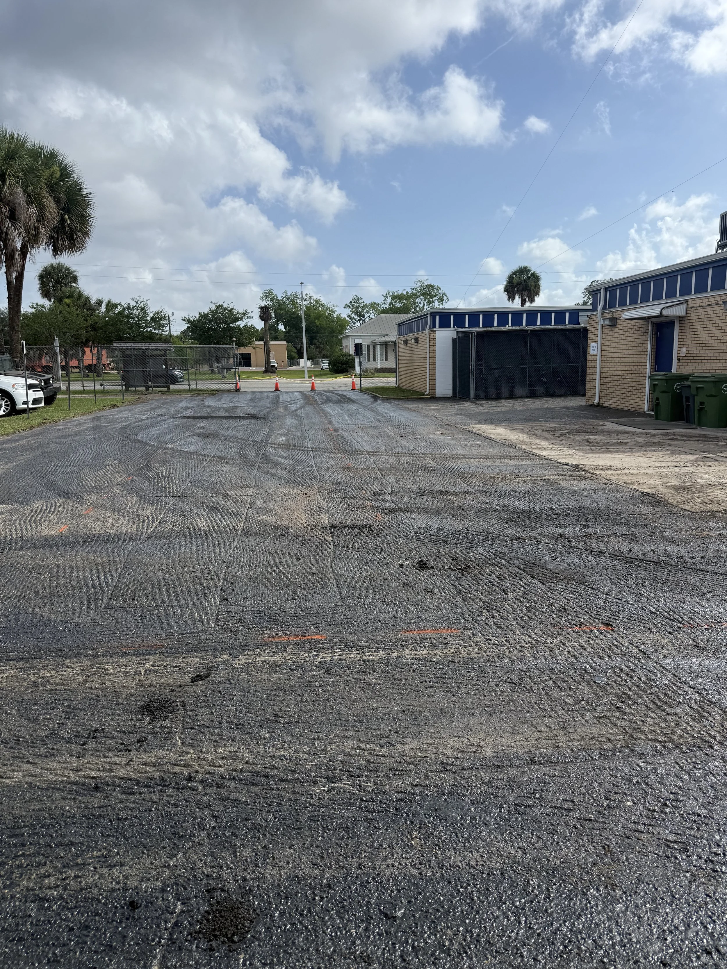 Newly paved parking lot with orange traffic cones blocking entrance, brick and white buildings, palm trees, and a partly cloudy sky.