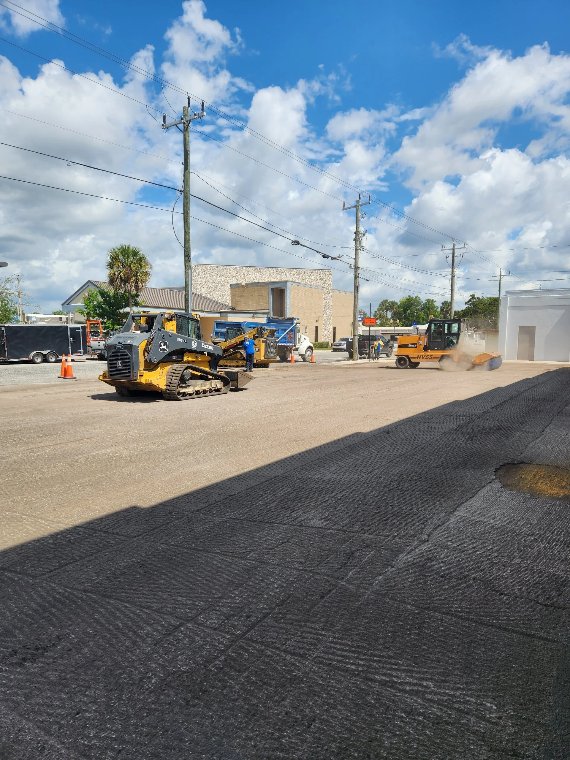 Construction worker operating a roller on a paved street, with other construction equipment and vehicles nearby, under a partly cloudy sky.