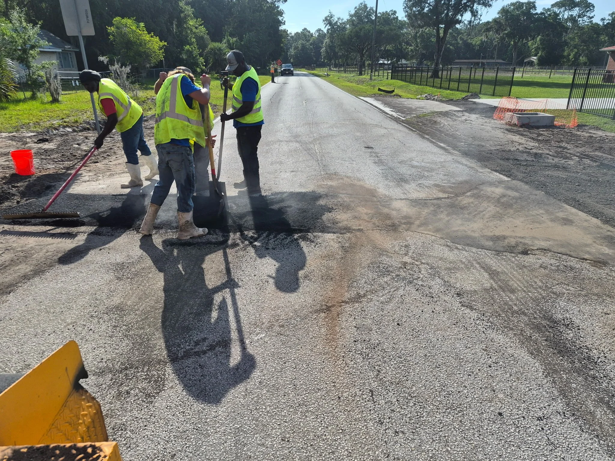 Workers in yellow safety vests and boots are repairing a road, digging and shoveling asphalt under a bright, sunny sky. A car is visible in the background, approaching from down the street, which is lined with trees and houses.
