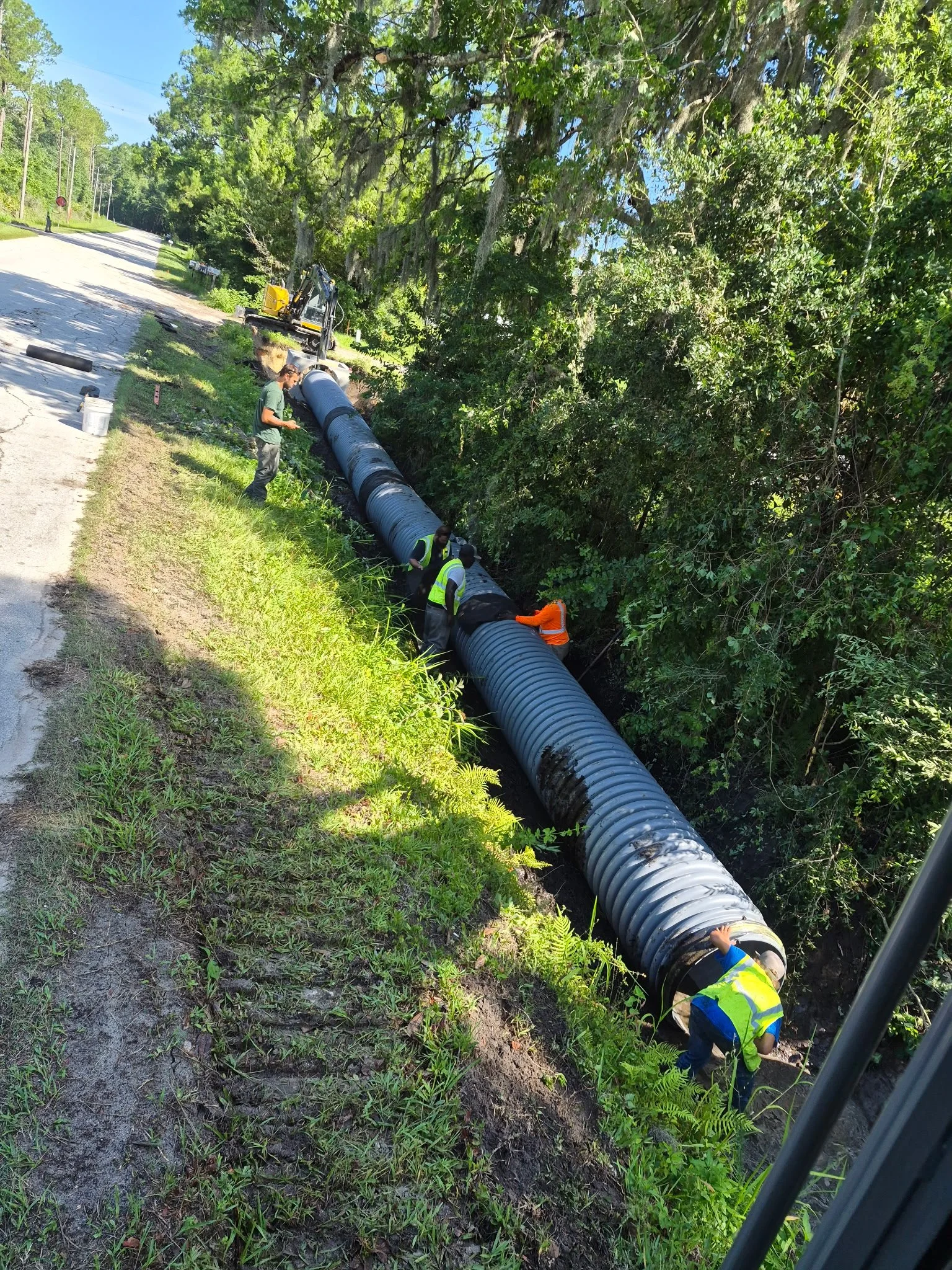 Workers in safety vests installing a large concrete drainage pipe along a roadside in a wooded area on a sunny day.