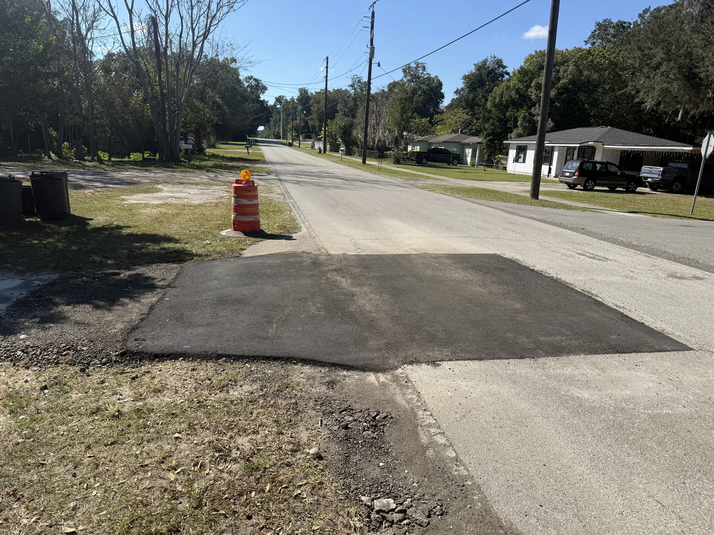A residential street with a construction barrel at the intersection. There are cars parked on the street, trees, and utility poles with power lines. The sky is clear with a few clouds.