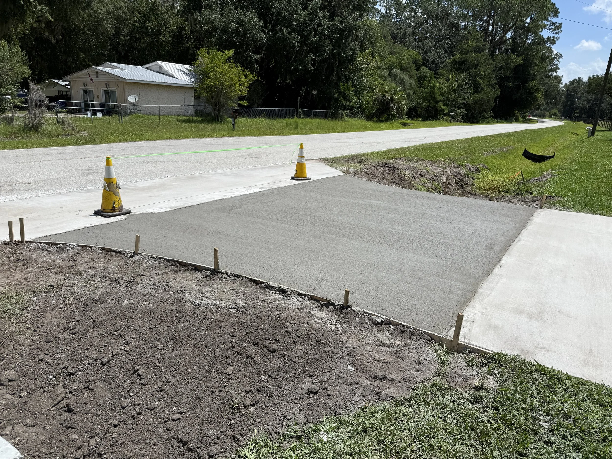 A residential street with a house, trees, and a driveway under construction. Two orange traffic cones and a green string mark a section of new concrete driveway. Construction materials and dirt are visible in the foreground.