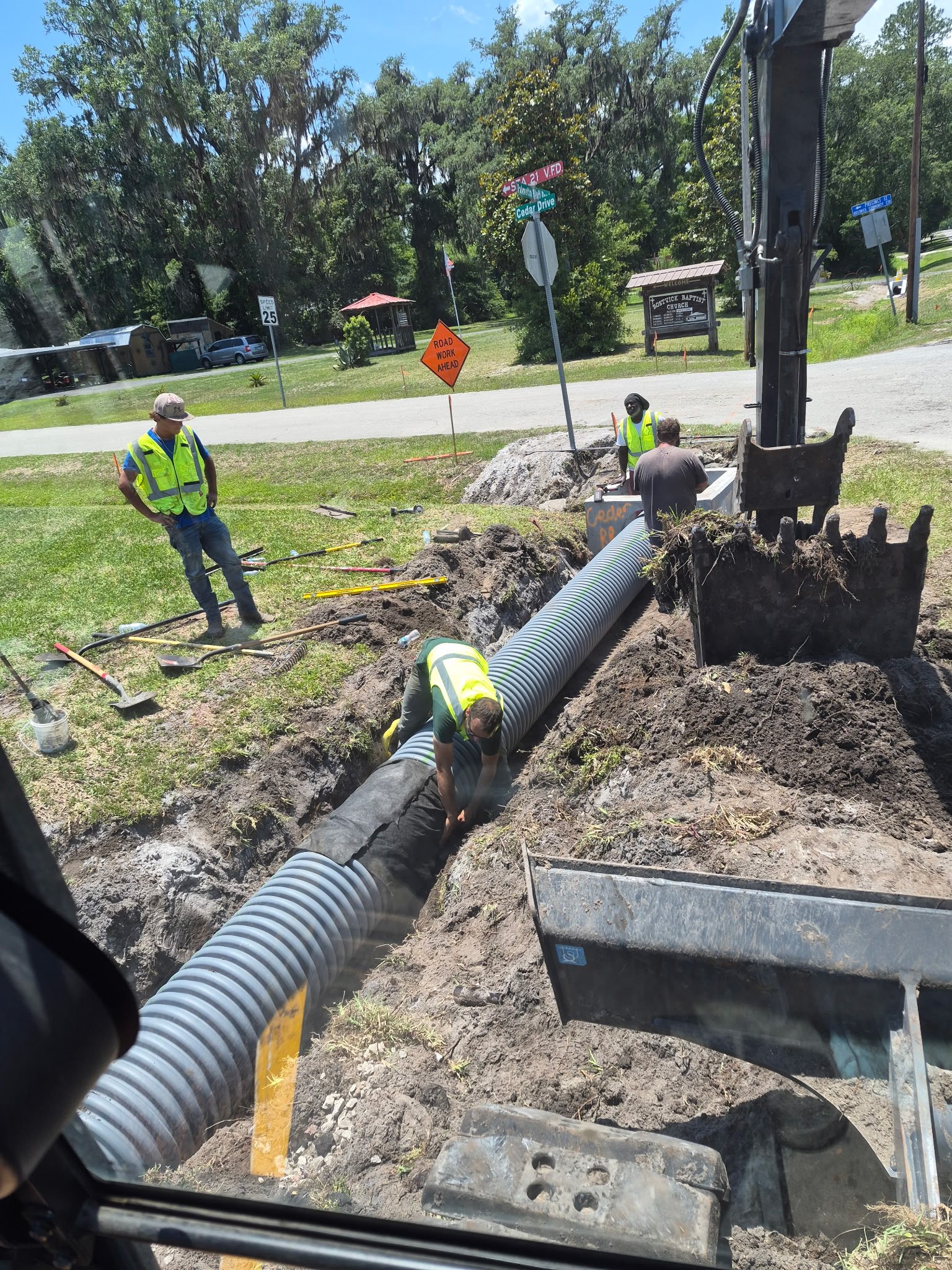 Workers installing large concrete drainage pipe in a construction site, with dirt and tools around, and trees and signs in the background.