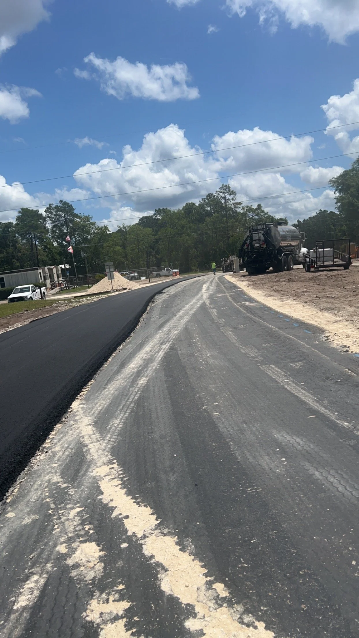 Construction site on a road with black asphalt and white gravel, construction vehicles, workers, and a partly cloudy sky.
