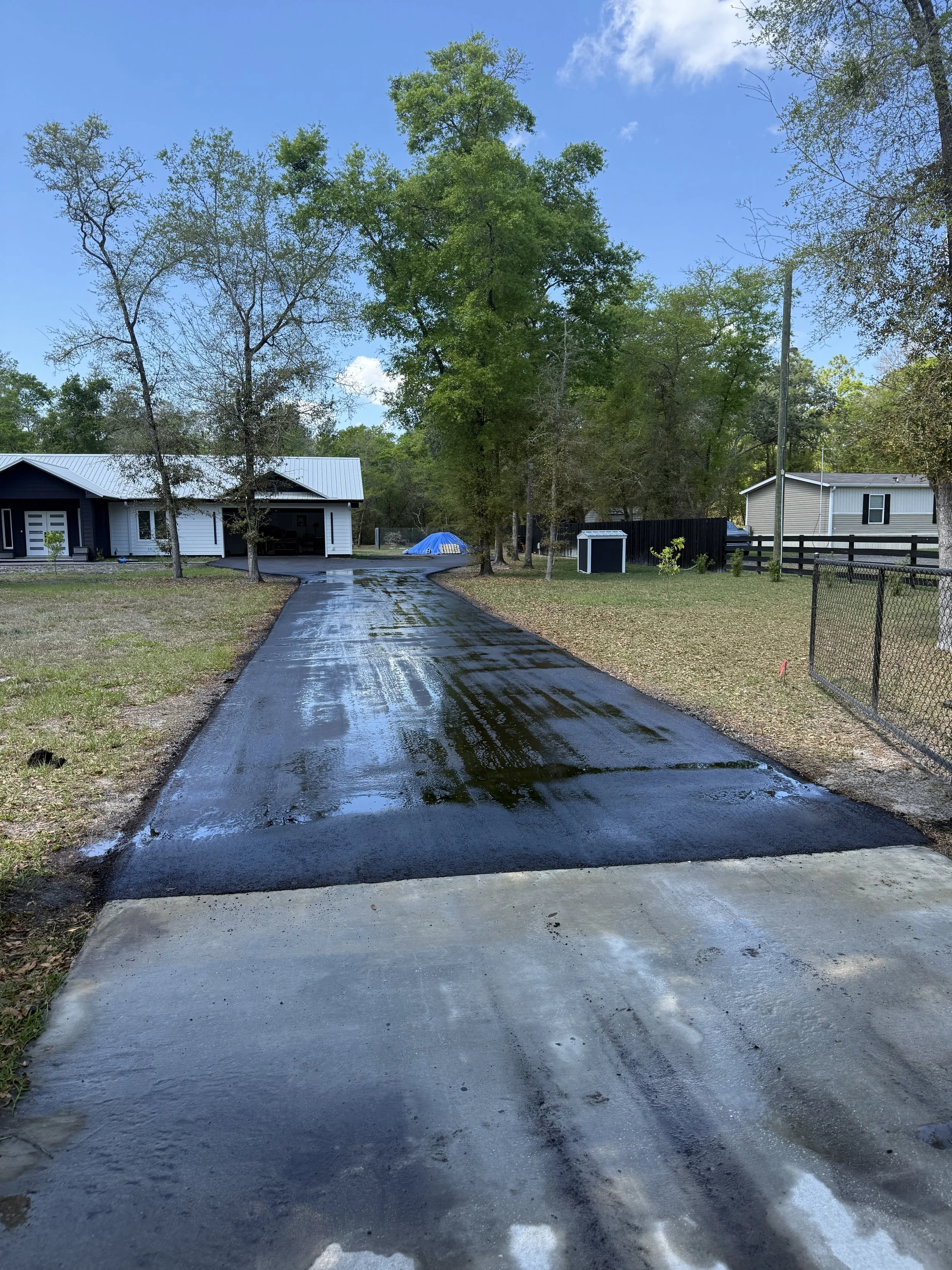 Freshly paved driveway with wet asphalt, leading to a house in a suburban neighborhood with trees, grass, and blue skies.