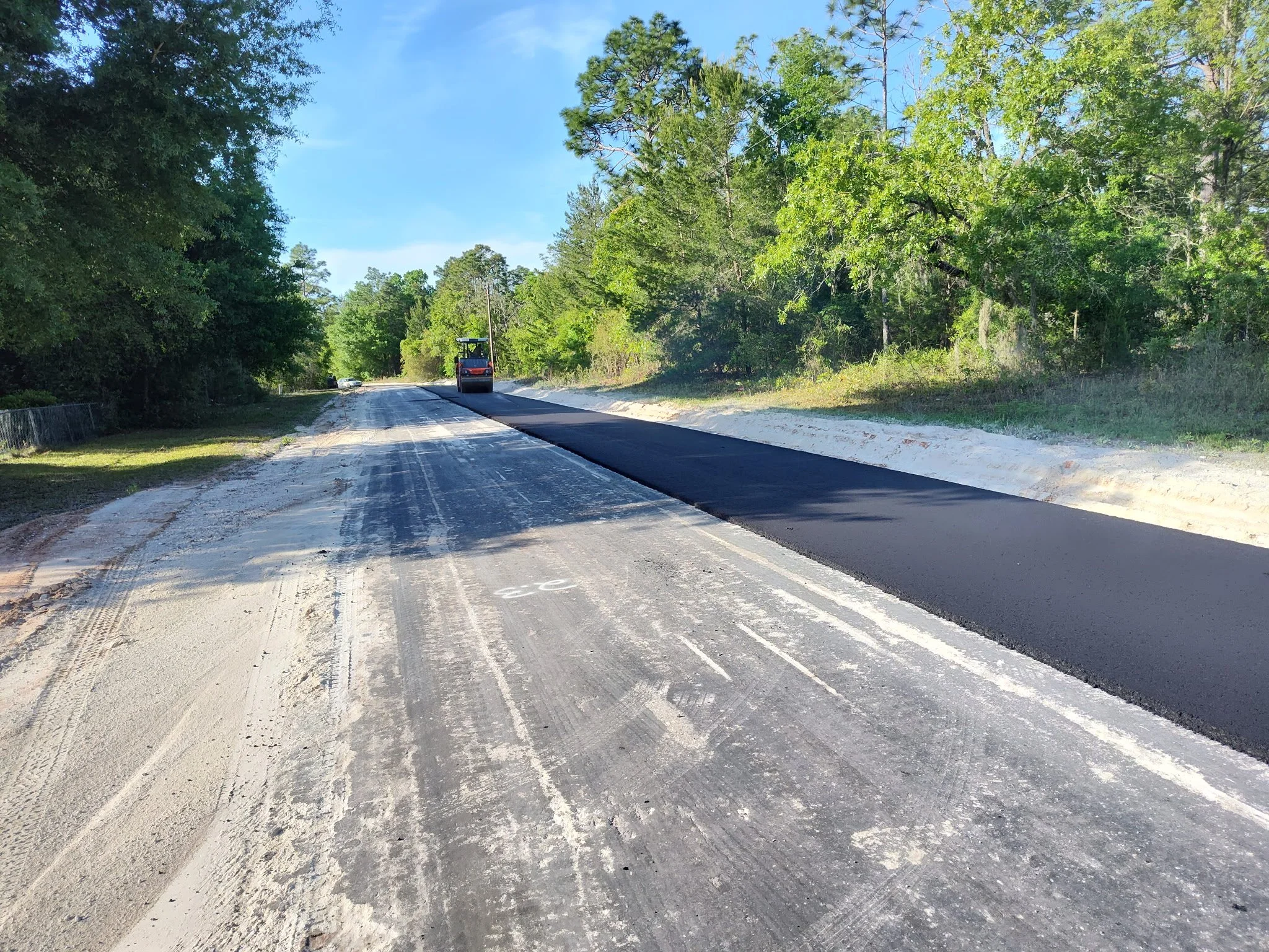 A rural road under construction with a paving machine laying asphalt, surrounded by green trees on both sides and a blue sky overhead.