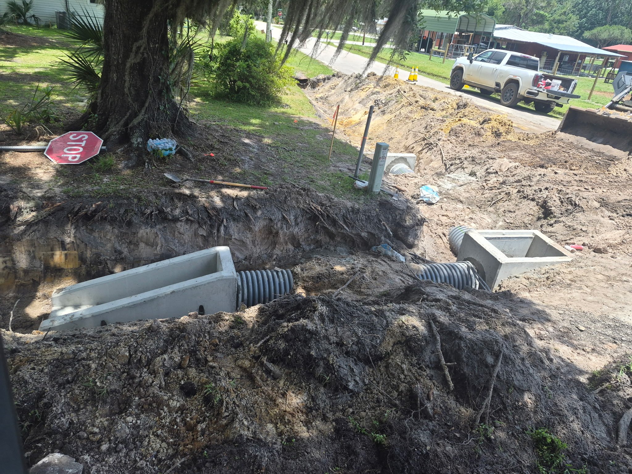 Construction site with a trench dug for underground concrete piping. A non-permanent stop sign is on the ground near a tree. Construction tools and cones are visible, along with a white pickup truck parked nearby.