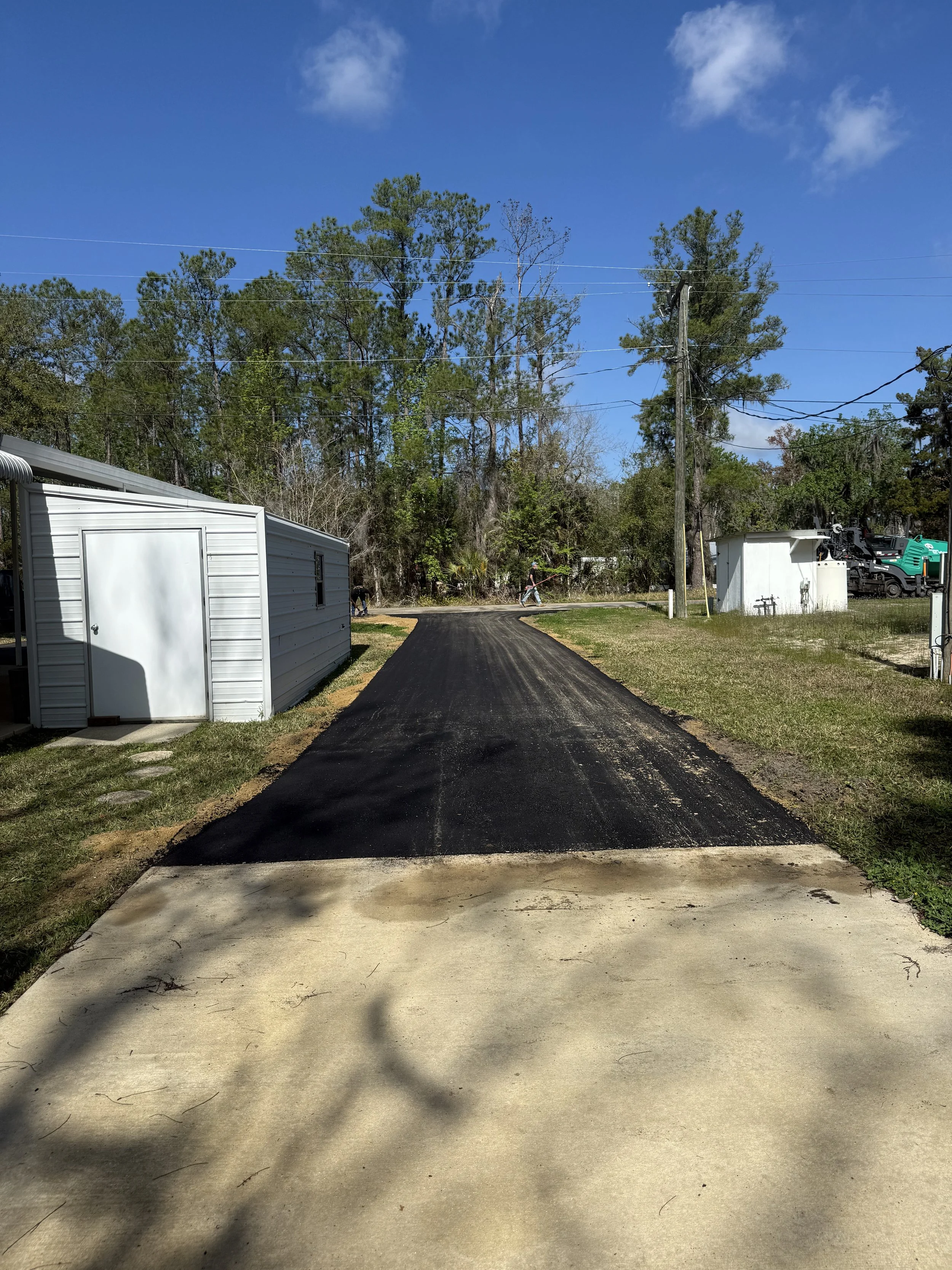 A newly paved blacktop driveway extends into a yard with a grassy lawn on each side, leading to a background of trees, power lines, and blue sky with a few clouds. There are structures on either side of the driveway, including a white shed and a mobi