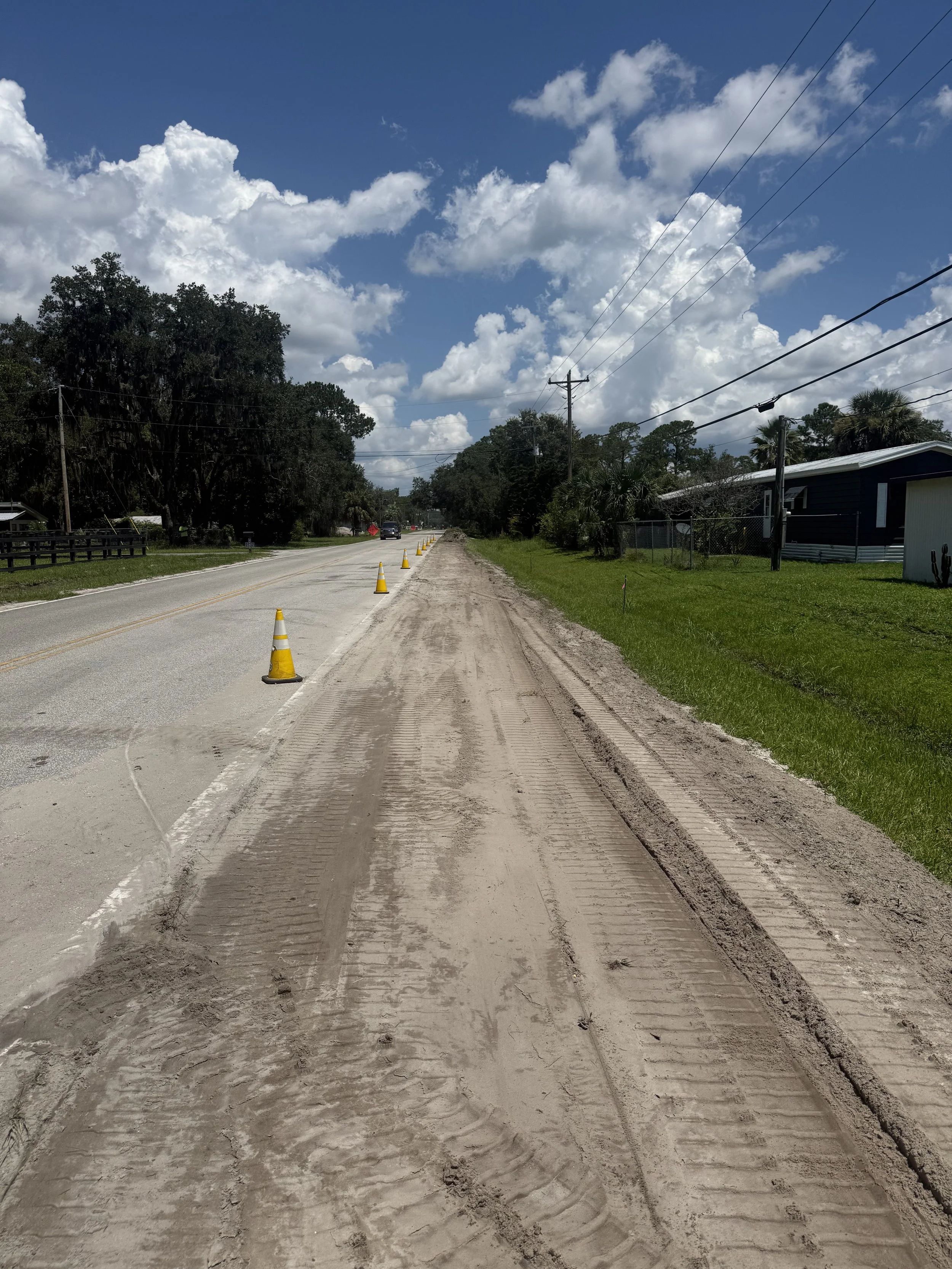 A rural road under a partly cloudy sky with yellow traffic cones, with a car and trees in the background and a residential house on the right side.
