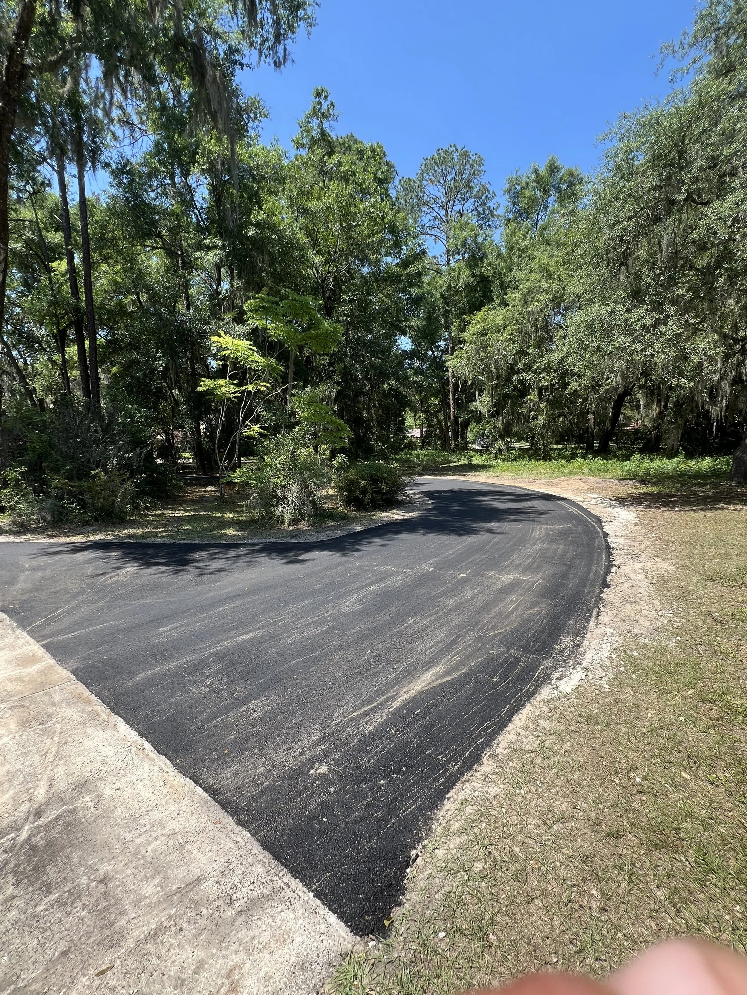 A new paved black asphalt driveway curves through a wooded area with tall trees, green foliage, and a clear blue sky.