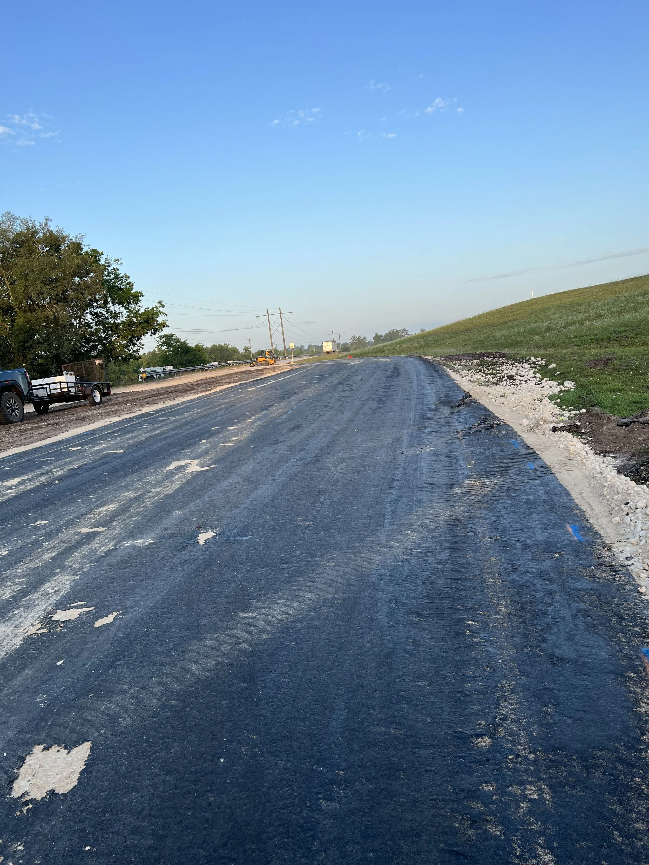 A road under construction with a vehicle on the side and construction equipment in the distance, surrounded by grassy fields and a clear blue sky.