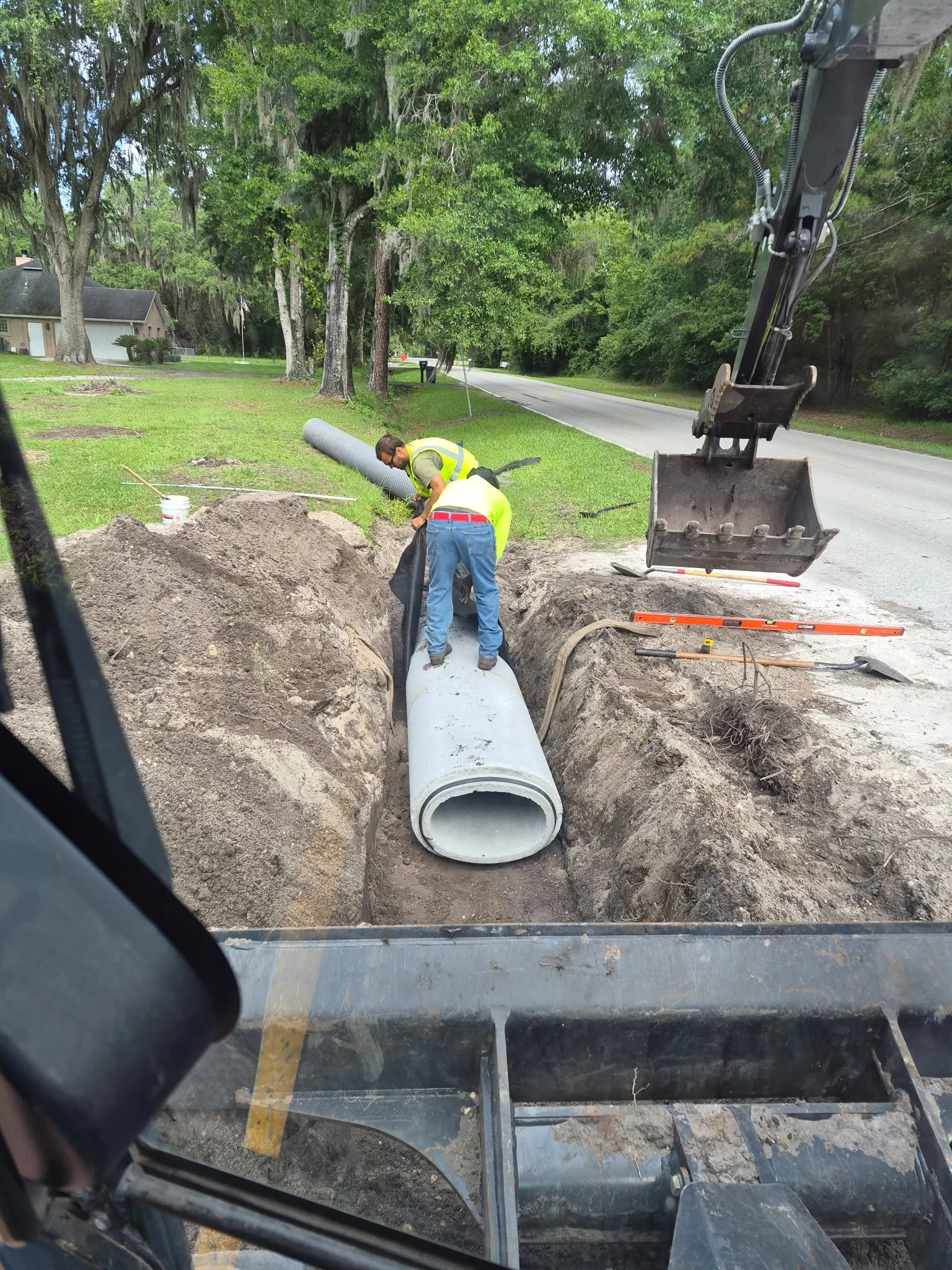 Construction workers installing a large concrete pipe in a dug trench on a residential street, with trees and houses in the background.
