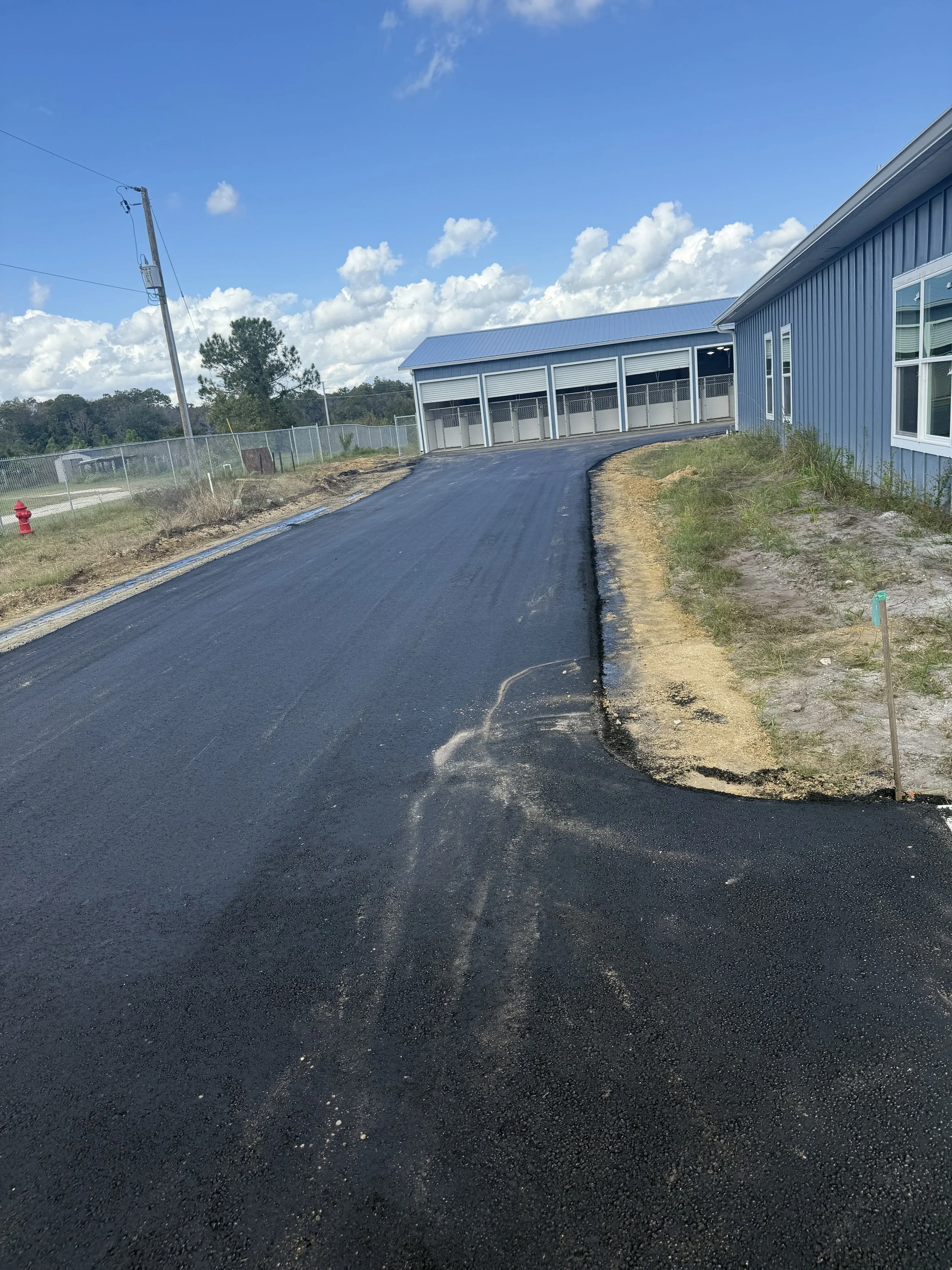 A newly paved blacktop road curves past a blue building with white trim, under a partly cloudy sky.