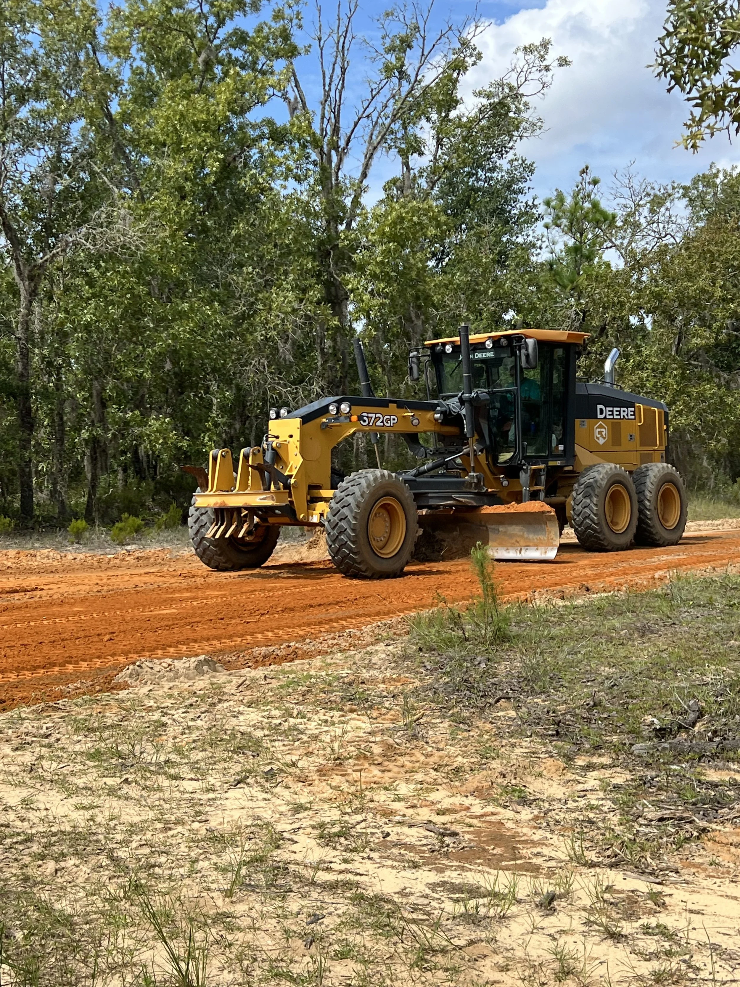 A yellow John Deere motorgrader working on a dirt road surrounded by trees and cloudy sky.