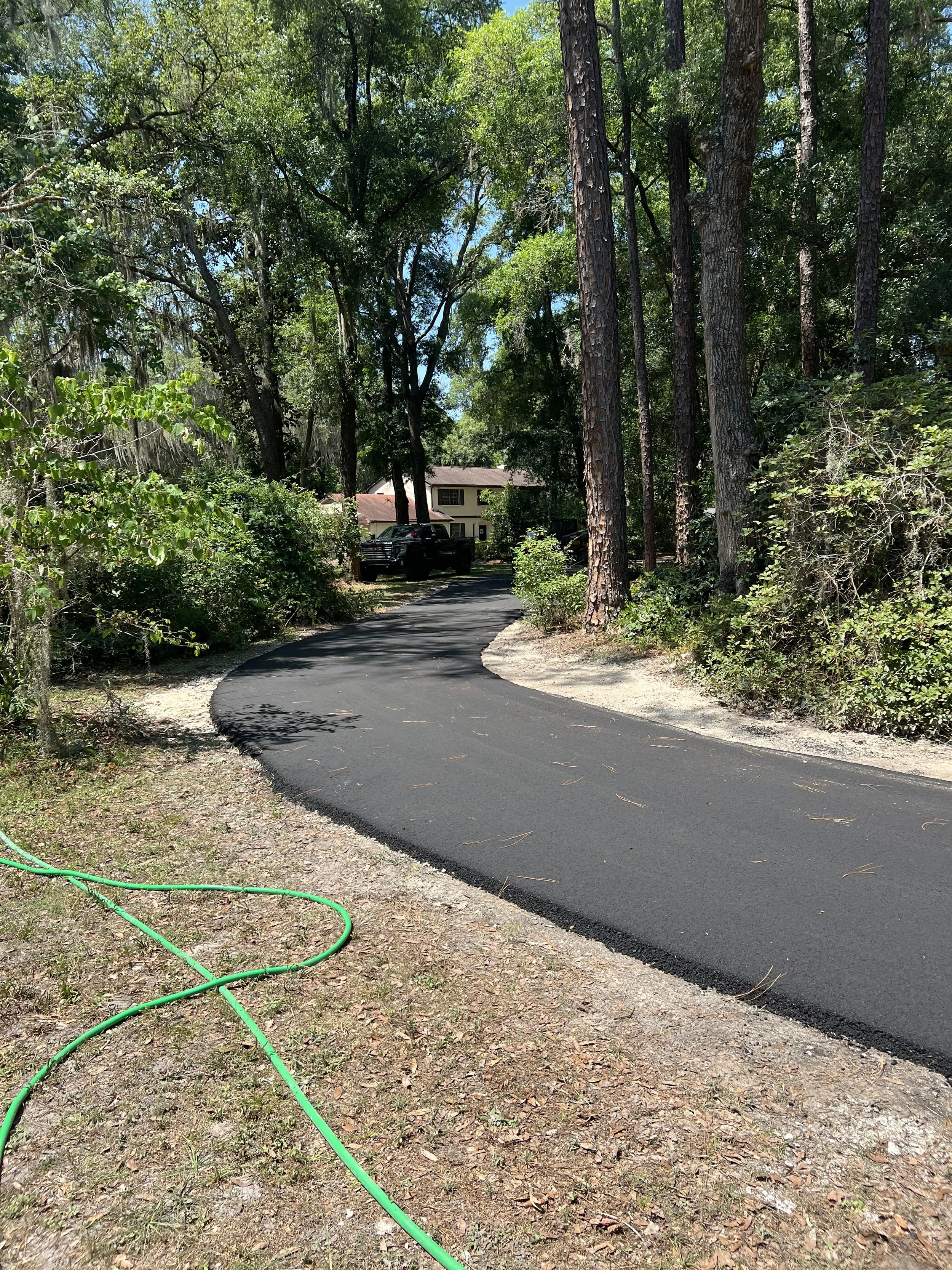 A newly paved black asphalt driveway curves through a wooded area with tall trees and green foliage, leading to a house in the background with a beige exterior and a red roof. A green garden hose lies on the ground near the edge of the driveway.