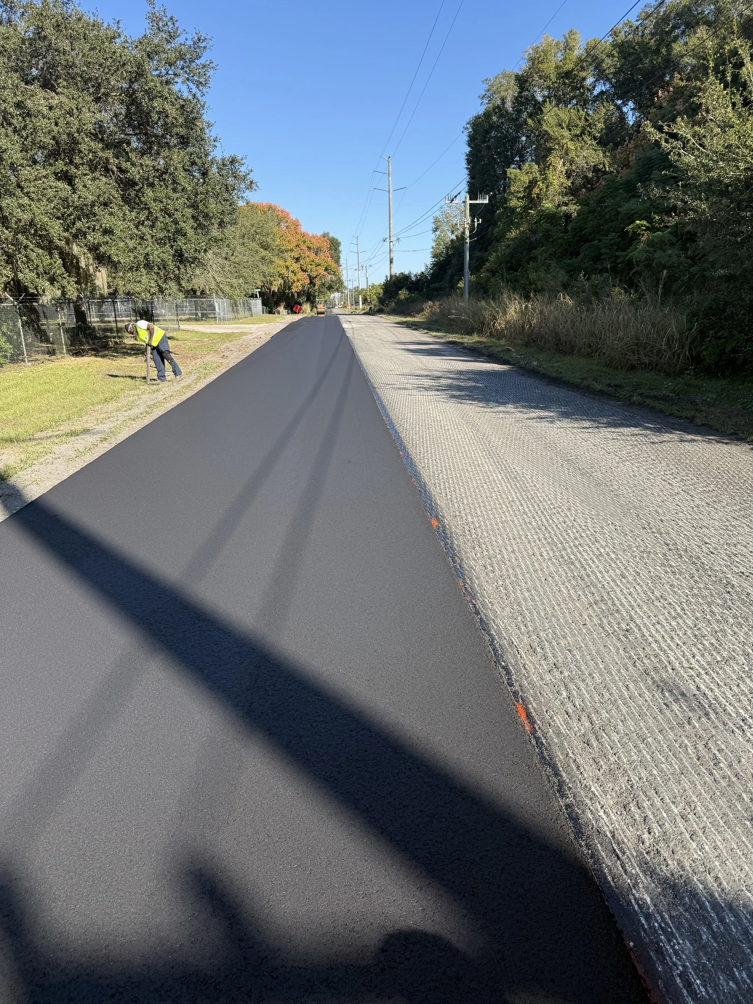 A worker in a yellow safety vest and helmet walking along the edge of a paved street with fresh black asphalt on a sunny day, with trees and power lines along the road.