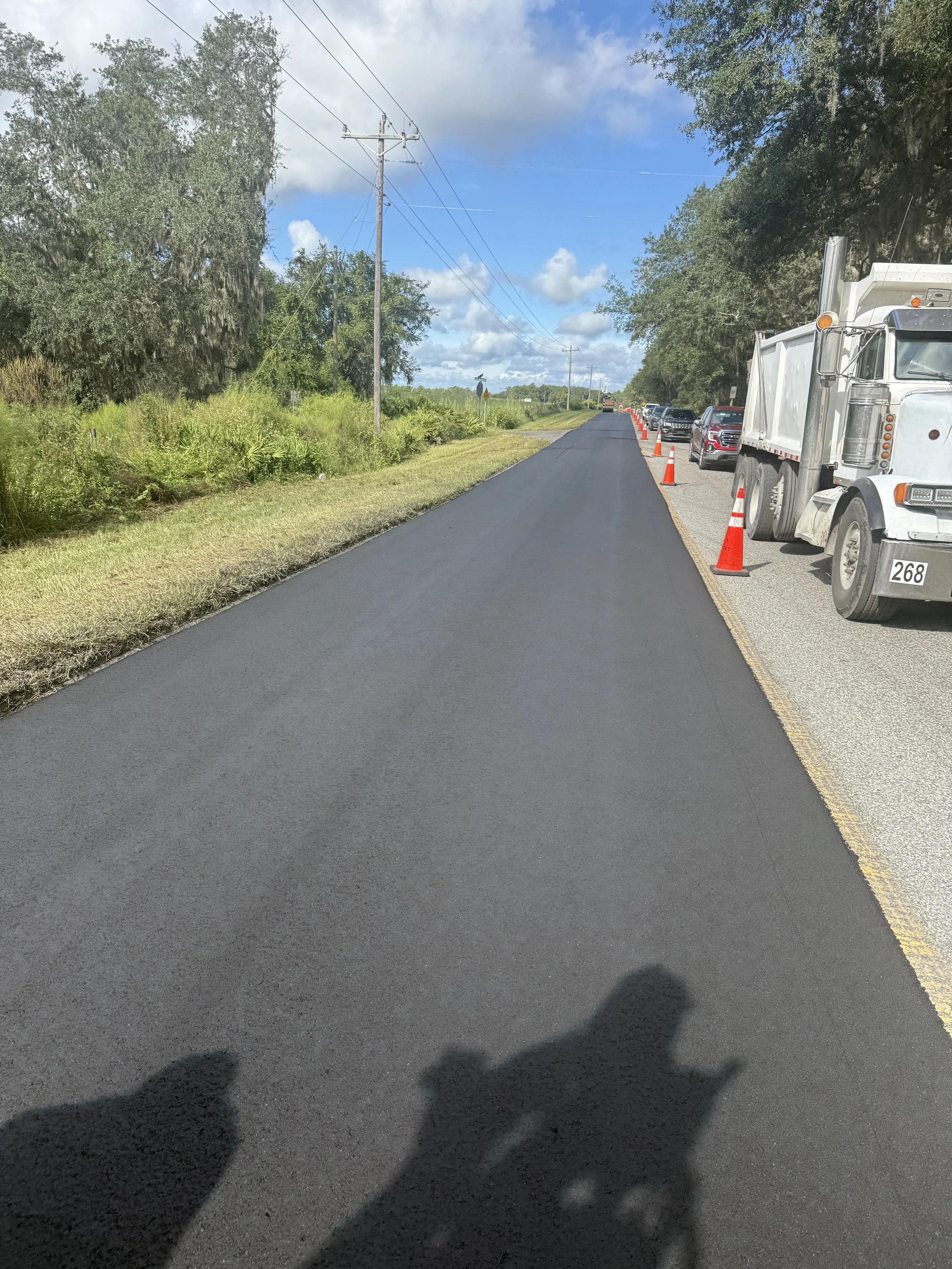 Newly paved black asphalt road with traffic cones and parked vehicles on the side, flanked by green vegetation and trees, under a partly cloudy sky.