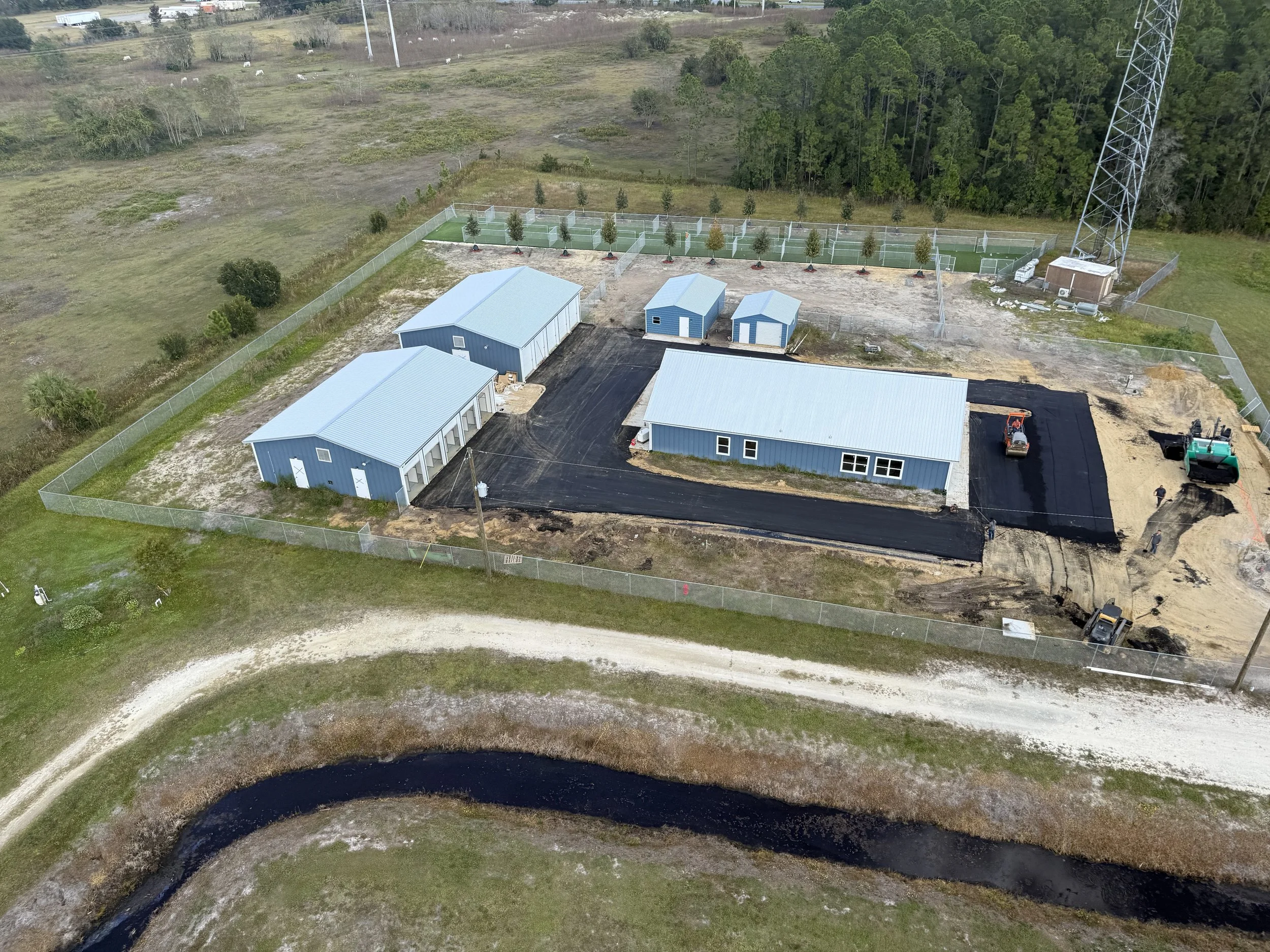 Aerial view of a construction site featuring large blue metal buildings, two smaller blue sheds, and an asphalt parking lot. Fencing surrounds a landscaped area with trees and a tennis court. Construction vehicles and workers are visible.