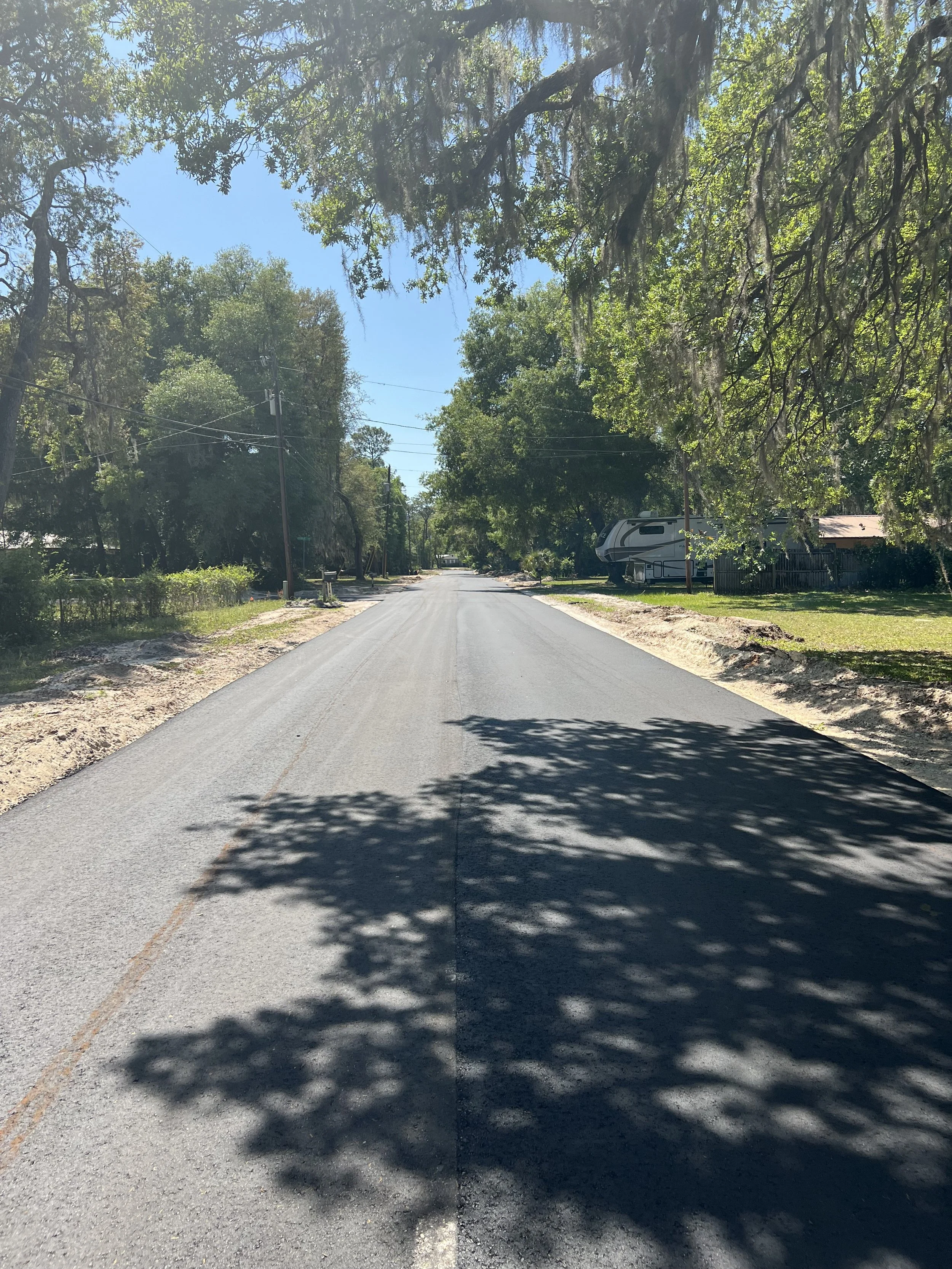 A paved rural street lined with trees, with some houses and a recreational vehicle (RV) visible on the right side, under a clear blue sky.